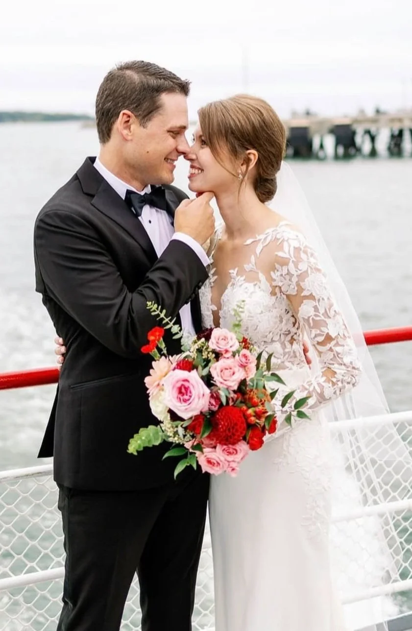 A bride and groom share a close moment on a boat, with water and a pier in the background. The bride holds a colorful bouquet, and the groom is wearing a black tuxedo.