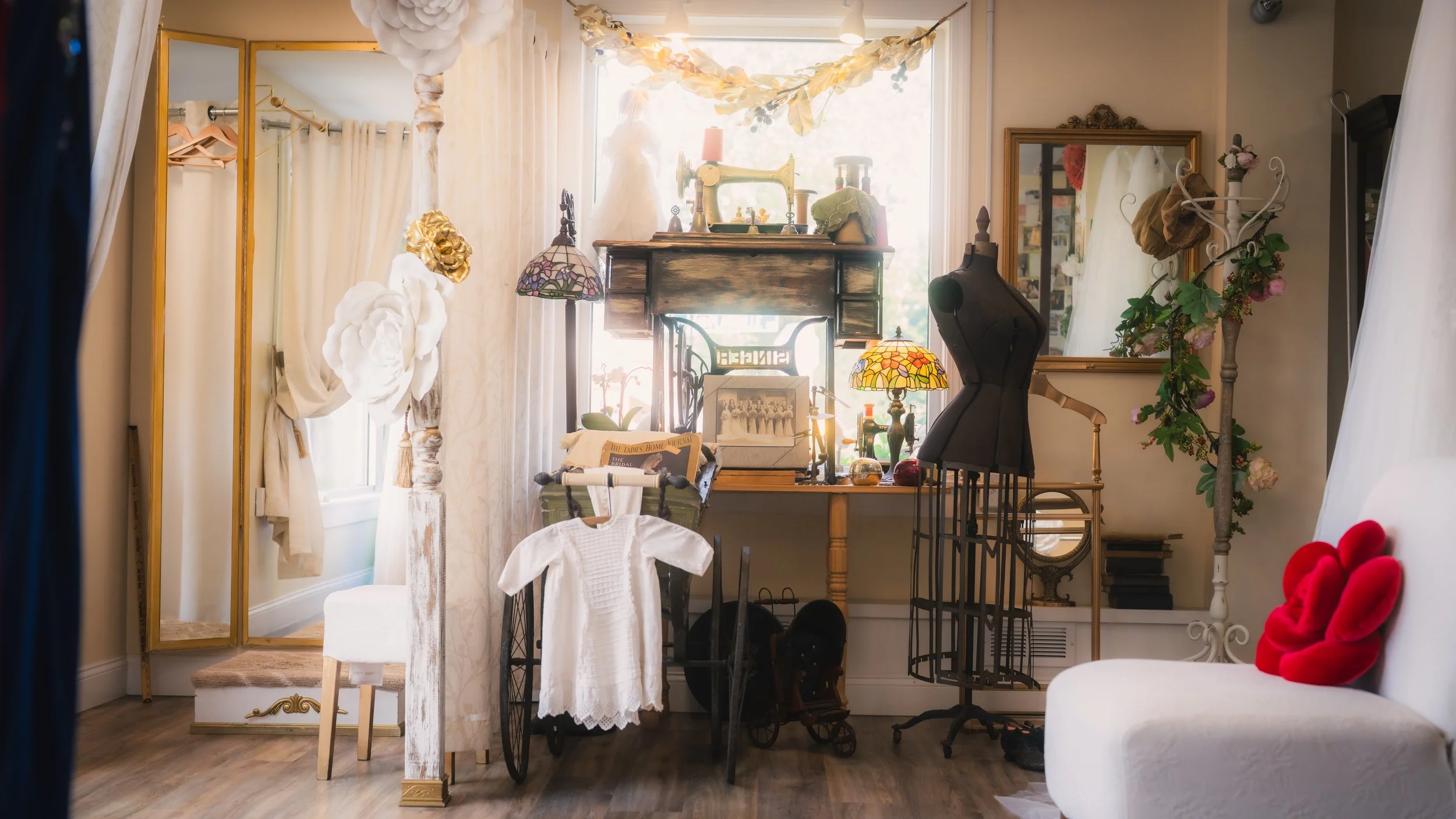 A vintage-style dressing room filled with various antique and decorative items, including a dress form, stained glass lamps, a mirror, a small bicycle, and a plush red footed chair.