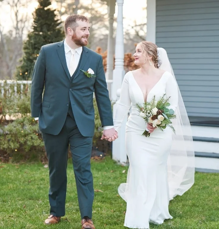 A bride and groom walking hand in hand outdoors, dressed in wedding attire. The bride is holding a bouquet of flowers and wearing a white dress with a veil, while the groom is in a dark suit with a tie. They are smiling and looking at each other, with trees and a house in the background.