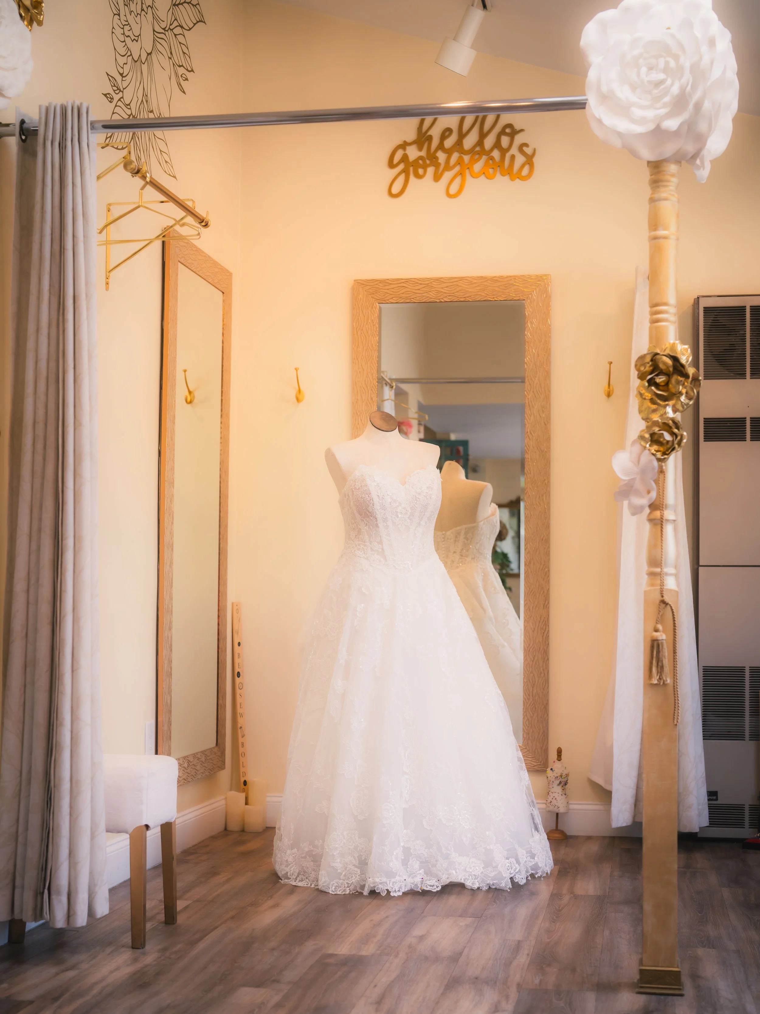 Wedding dress displayed on a mannequin in a boutique, with a mirror behind it, a decorative gold sign that says 'hello gorgeous' above, and a floral lamp on the right side.