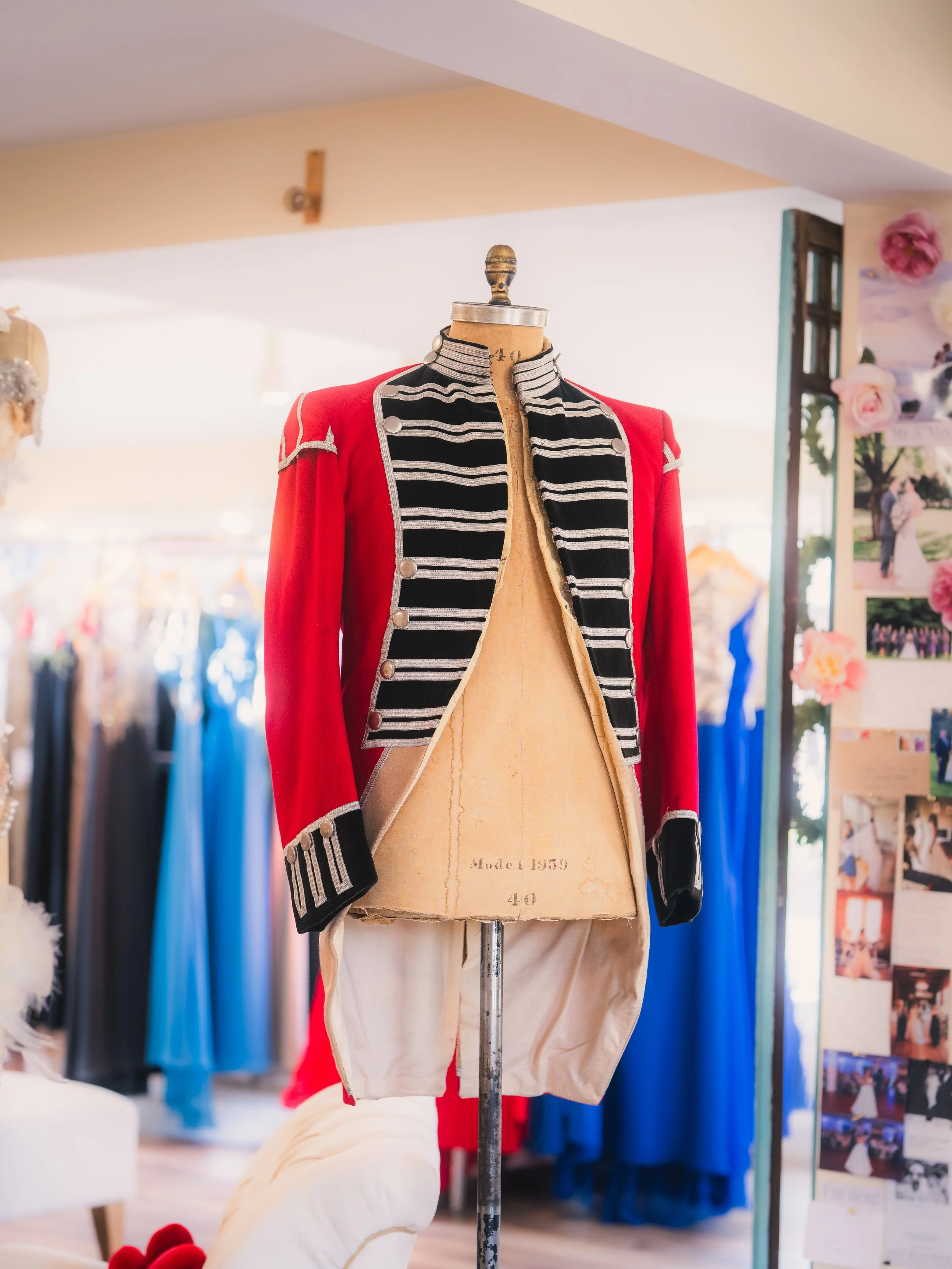A vintage military-style jacket displayed on a dress form in a boutique or costume shop, with racks of dresses and photos in the background.