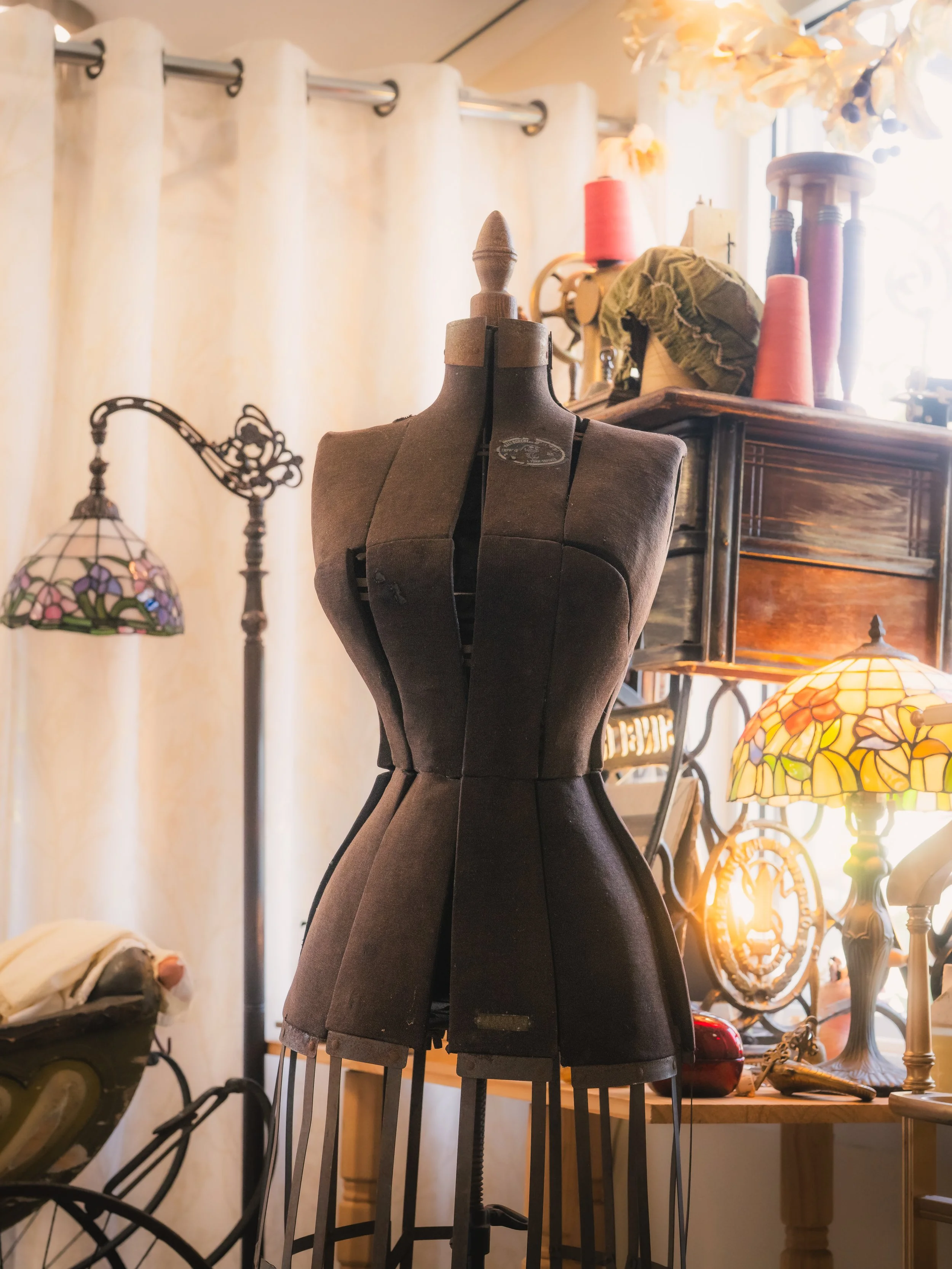 A vintage dress form mannequin in a room filled with sewing and crafting supplies, including lamps, spools of thread, and fabric.