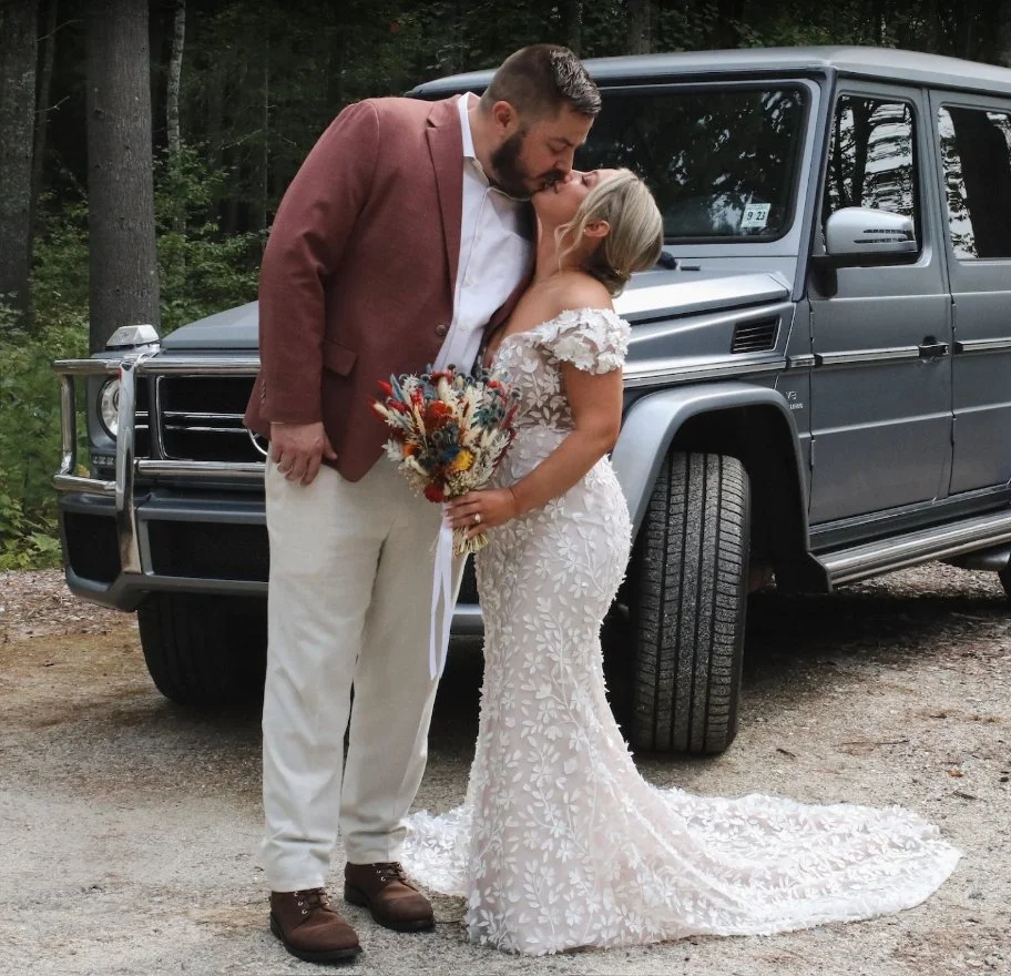 A newlywed couple sharing a kiss in front of a gray SUV in a wooded outdoor setting. The bride wears a lace wedding gown and holds a bouquet of flowers; the groom wears a brown blazer, white shirt, and white pants.