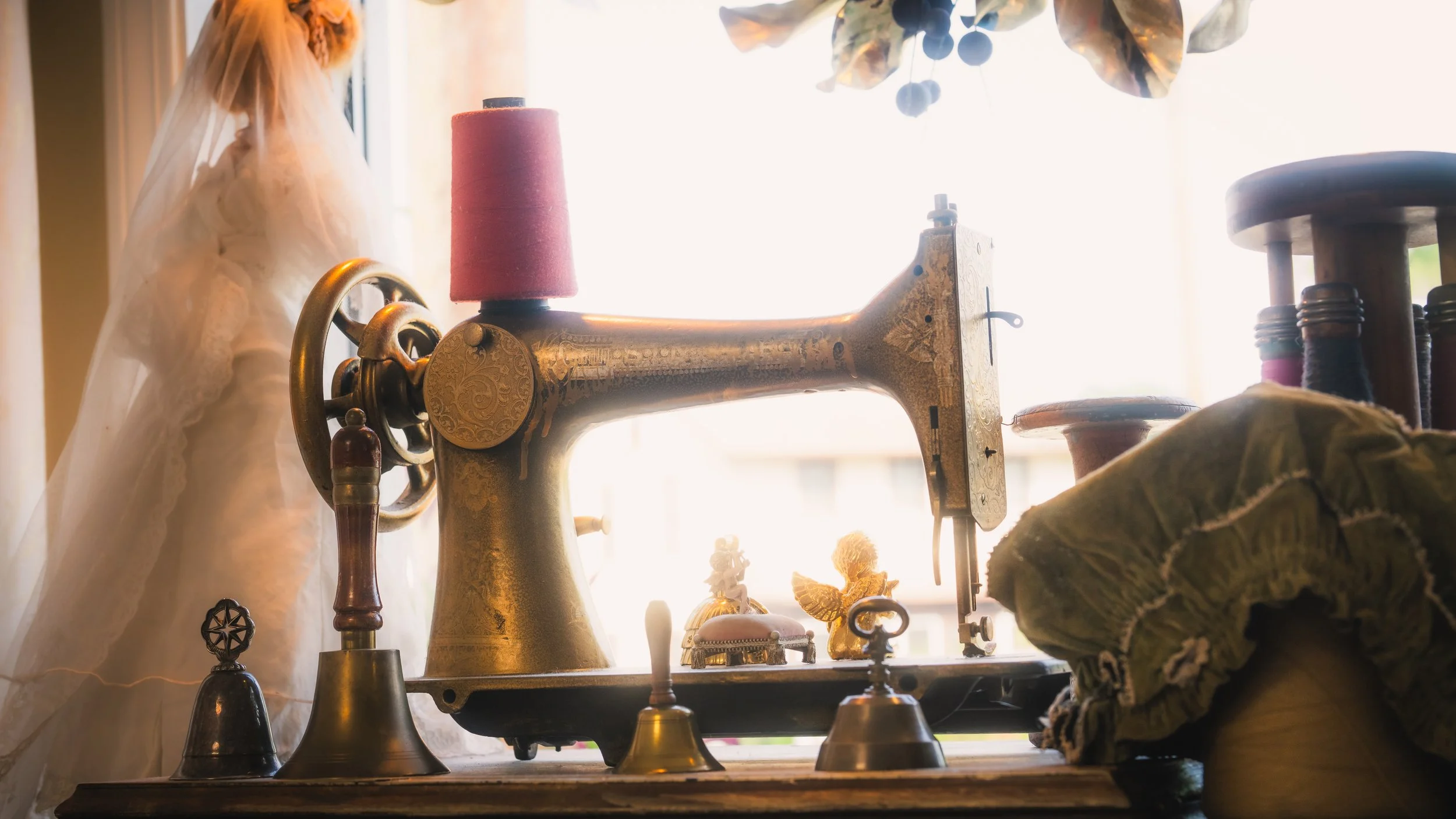 Vintage gold-colored sewing machine on a wooden table with sewing accessories, decorative figurines, and a fabric piece, illuminated by natural light coming from a nearby window.