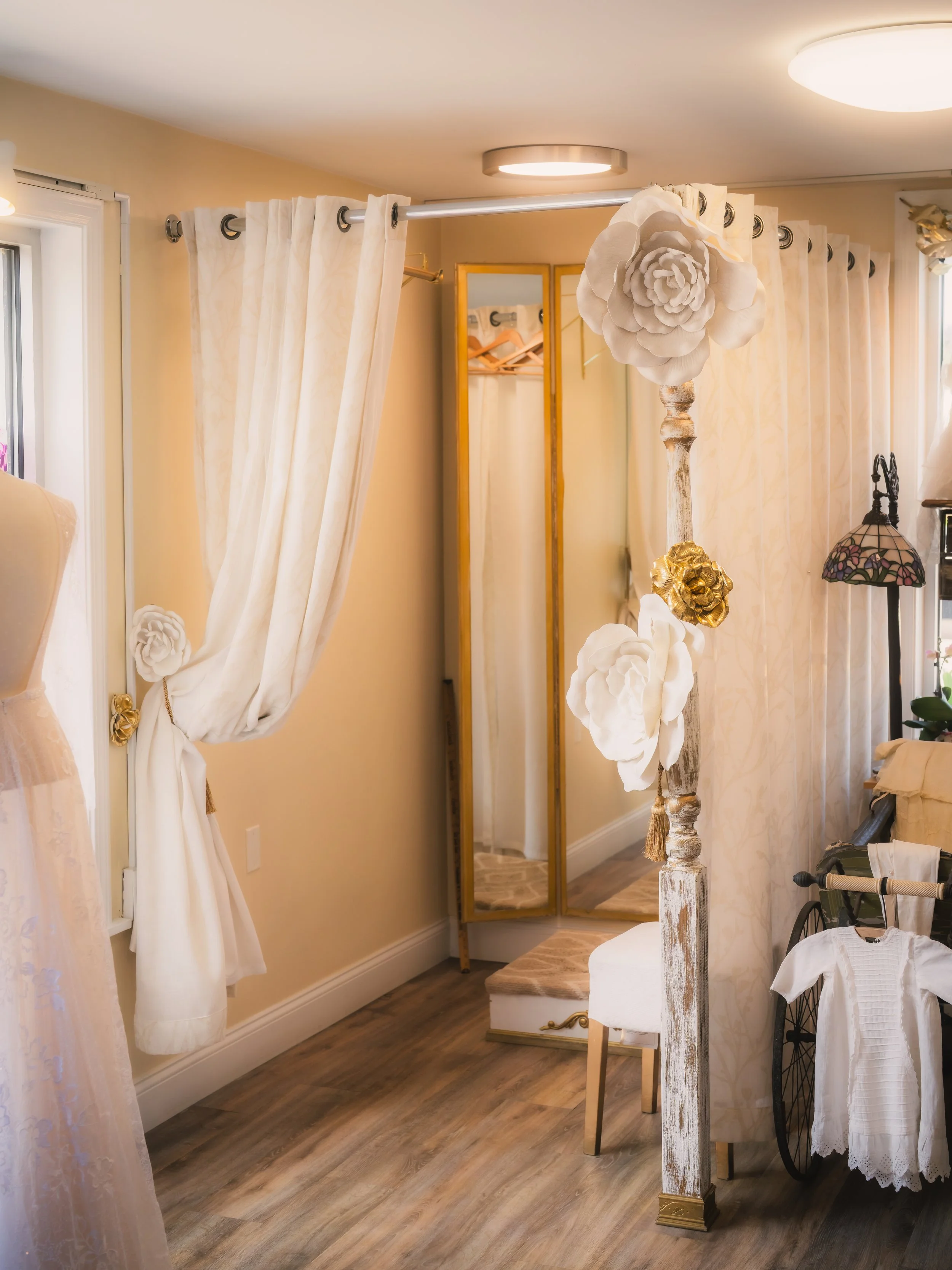 A cozy room with cream-colored curtains, a tall mirror, and floral decorations, including large paper flowers hanging from a distressed white wooden post. A small upholstered stool and a vintage-style lamp are also visible.