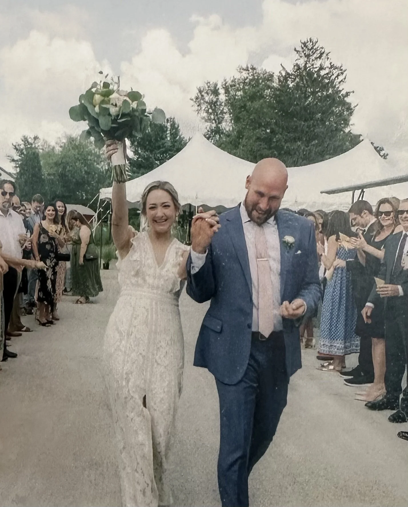 A bride and groom celebrating at their wedding reception outdoors, with the bride raising a bouquet and smiling while the groom appears to be emotional, as guests stand under tents.