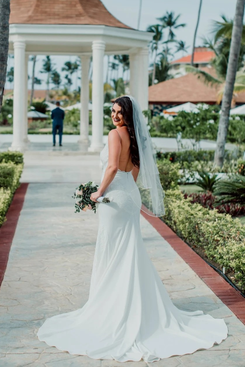 A bride in a white wedding dress with a long train and veil holding a bouquet, smiling on a garden pathway with a pavilion and palm trees in the background.