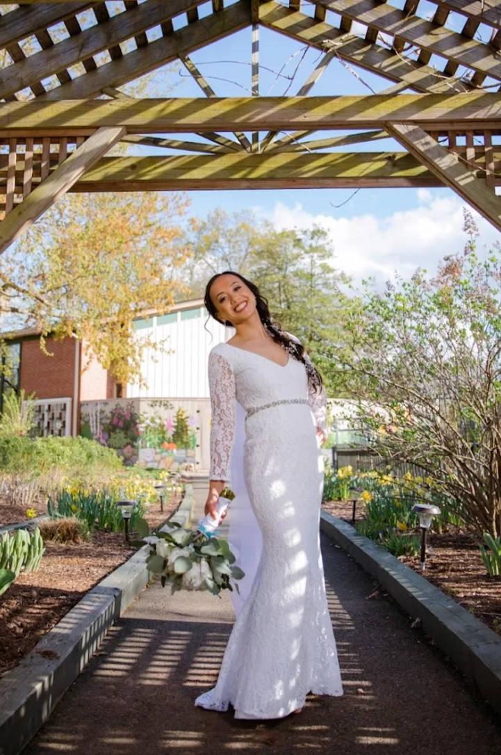 Woman in a white lace wedding dress holding a bouquet, standing on a garden path under wooden pergola, with trees and blooming plants in the background.