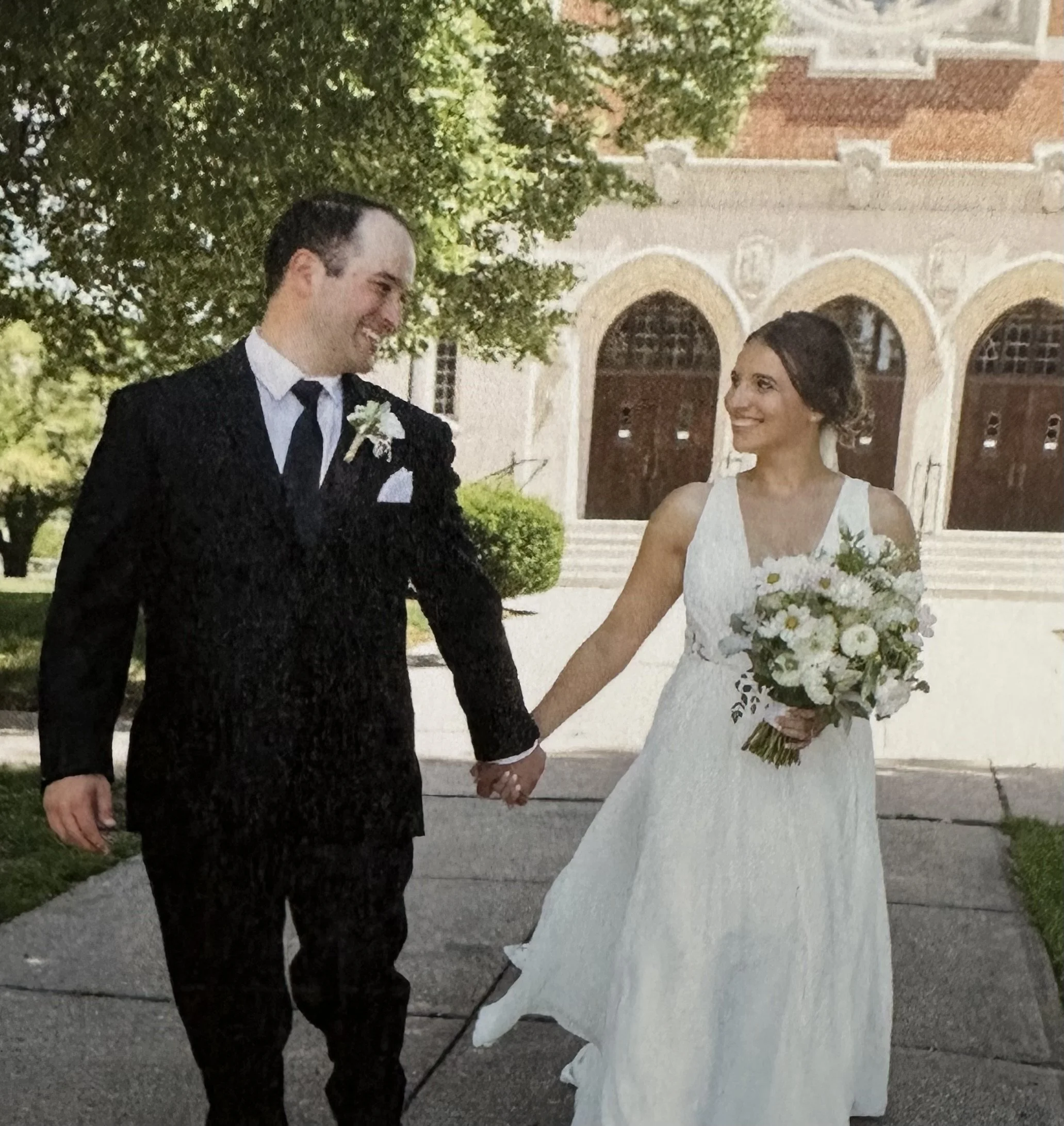 A bride and groom holding hands and smiling at each other outdoors on their wedding day. The bride is wearing a white wedding gown and holding a bouquet of white flowers, while the groom is dressed in a black tuxedo with a white shirt and tie. A historic building with arches is in the background.