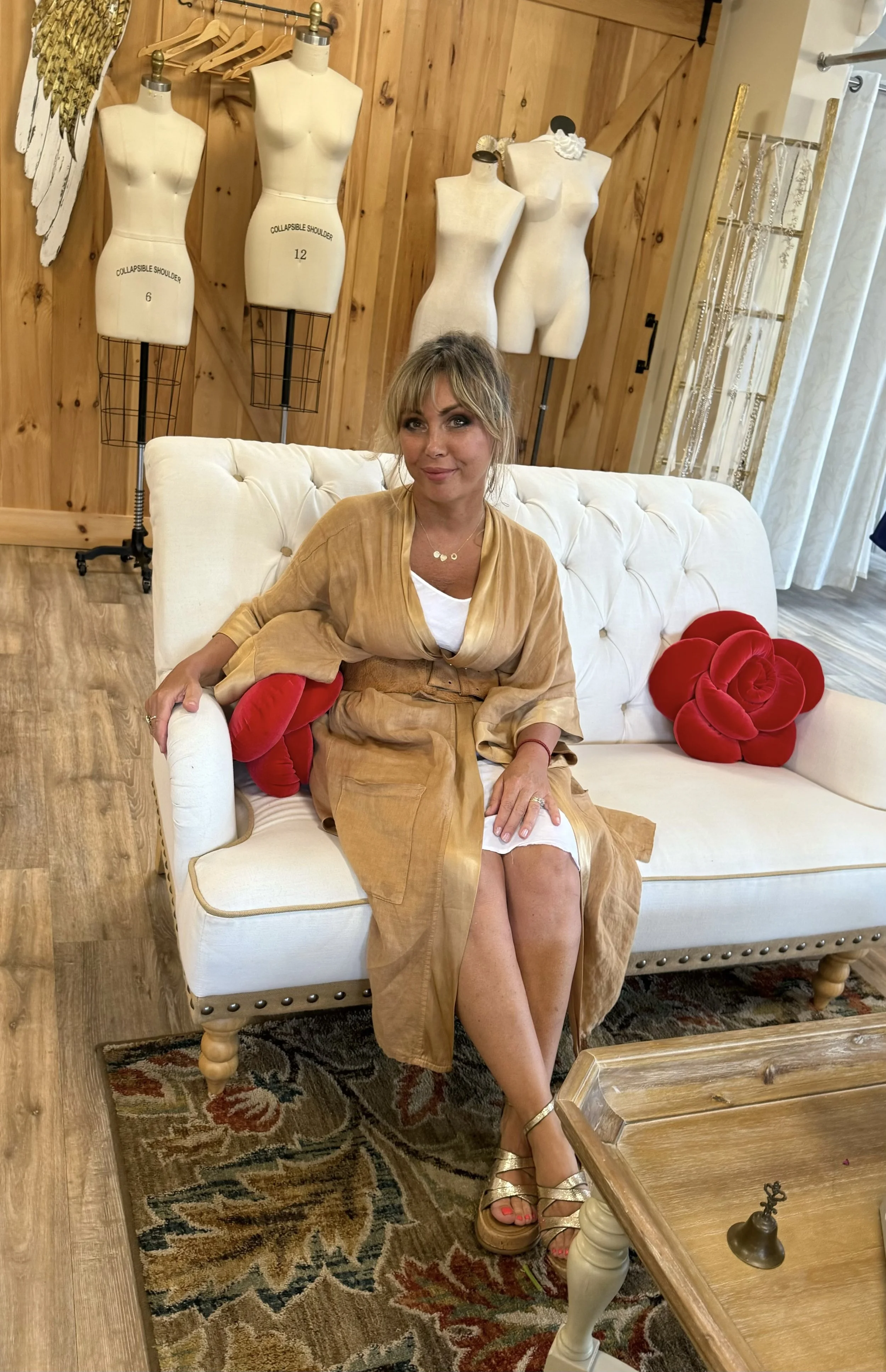 A woman sitting on a white tufted sofa with red plush rose pillows, in a room with a wood-paneled wall. The woman is wearing a tan dress and gold sandals, and the background features dress mannequins and jewelry displays.