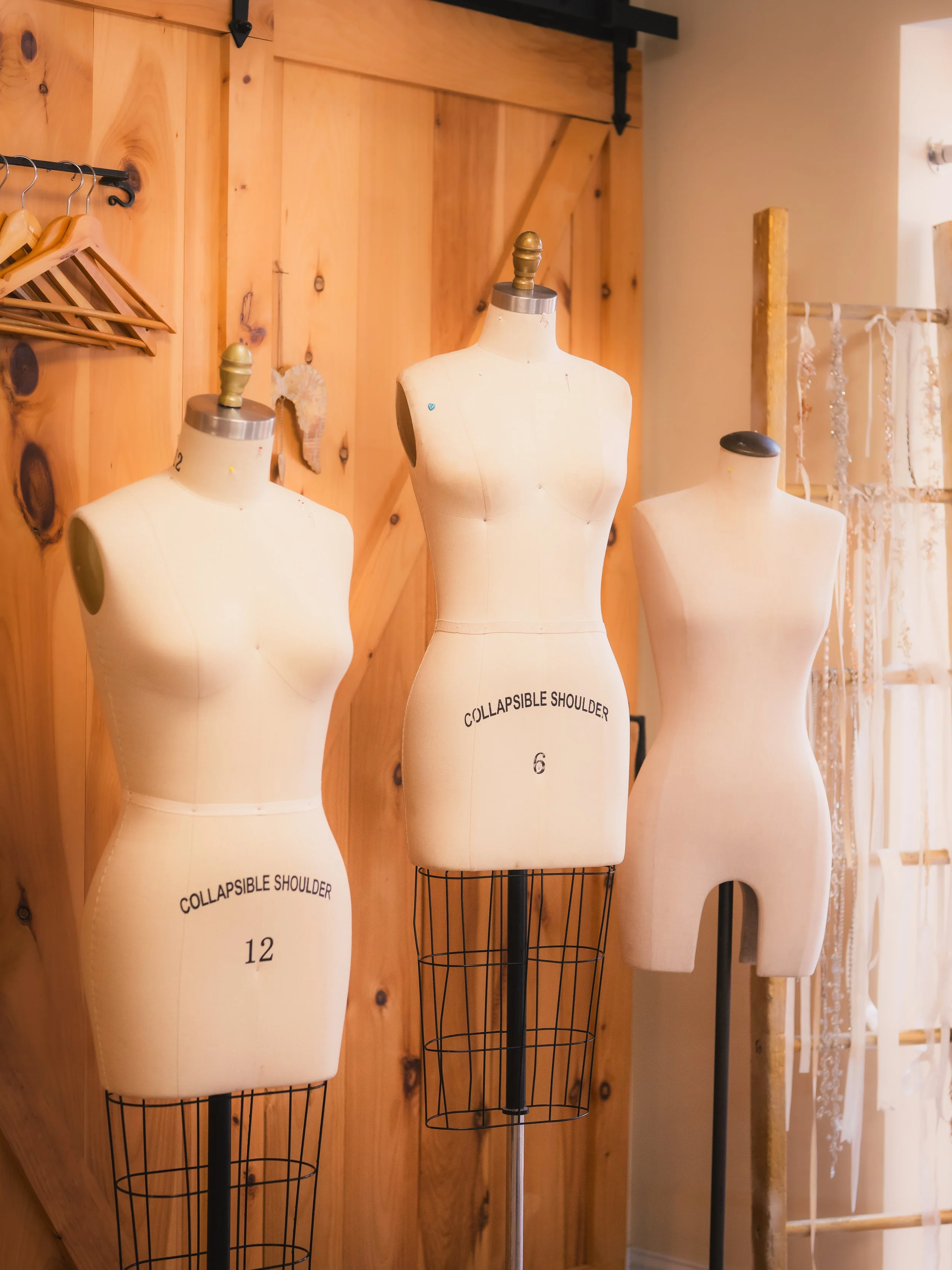Three dress form mannequins standing in a sewing or design studio, each labeled with different sizes, against a wooden wall backdrop.