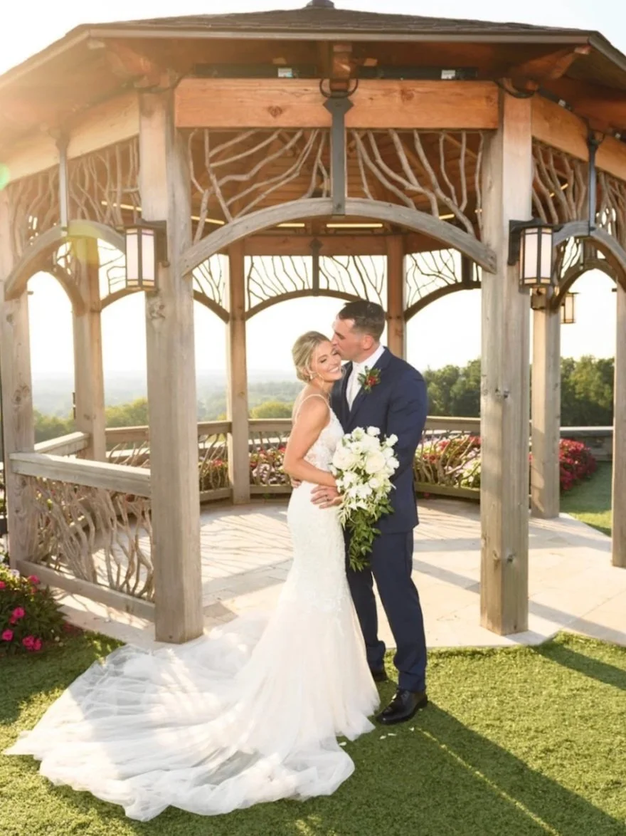 A newlywed couple standing under a wooden gazebo, embracing and smiling. The bride is wearing a white wedding gown and holding a large bouquet of white flowers, while the groom is in a navy blue suit. The background shows a scenic outdoor view with trees and a bright sky.