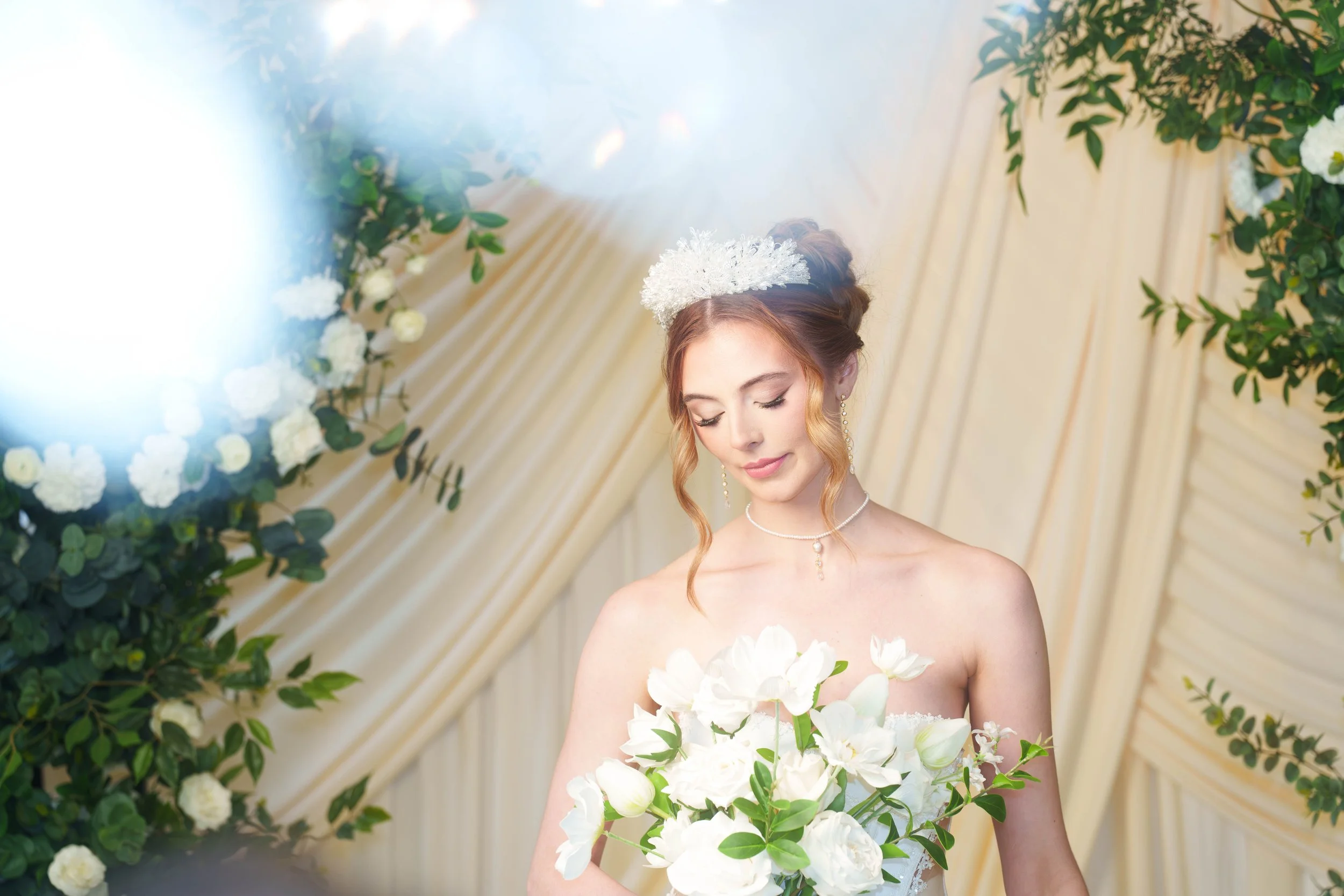 Soft bridal portrait of a woman in a white gown holding a white floral bouquet, framed by cream drapery, greenery, and bright chandelier bokeh in the foreground.