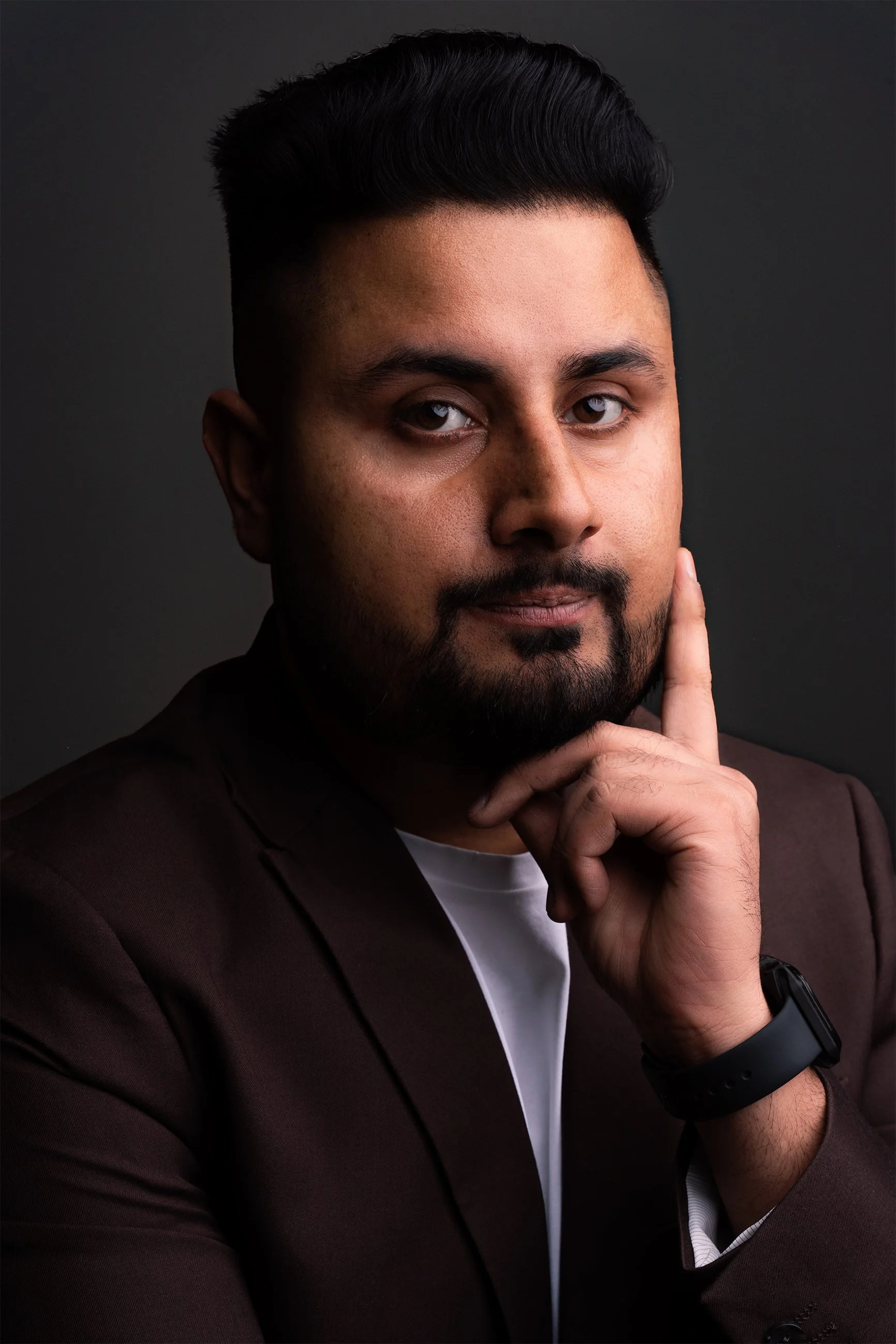 Studio portrait of a man in a dark blazer resting his hand on his face, lit with soft directional lighting against a neutral background.