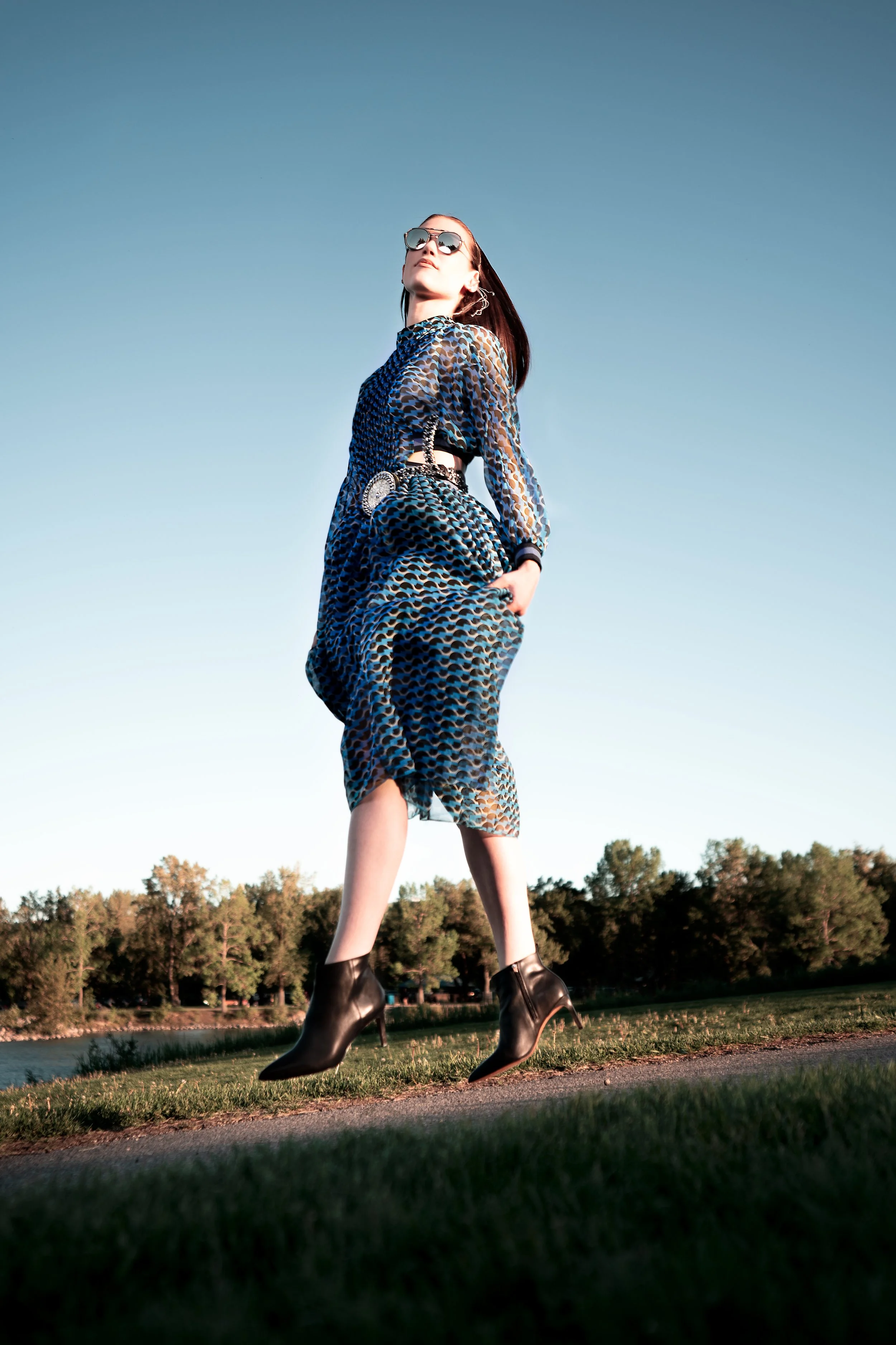 Fashion portrait of a woman in a patterned dress and black heeled boots jumping in mid-air outdoors, captured from a low angle with warm evening light and park landscape.
