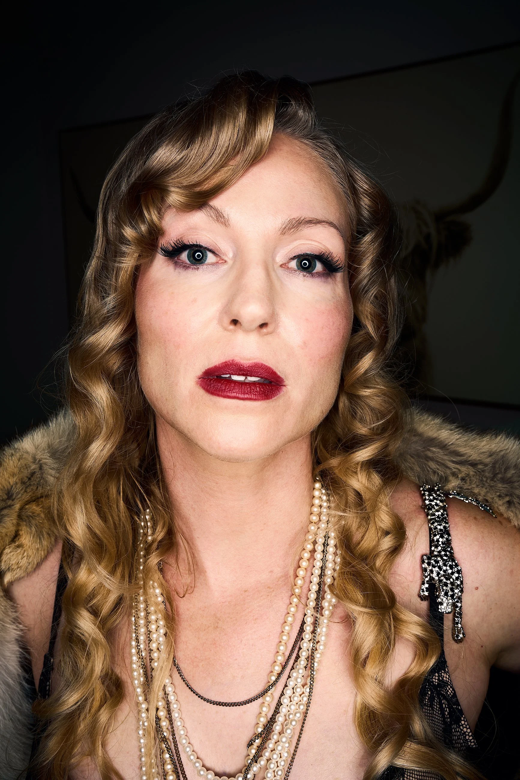 Vintage-inspired portrait of a woman with curled hair, red lipstick, and pearl necklaces in dramatic studio lighting.