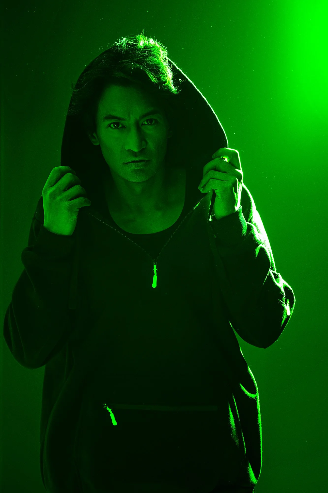 Cinematic low-key portrait of a man in a dark hoodie, gripping the hood as vivid green lighting cuts across the frame, creating a bold, high-contrast editorial look.