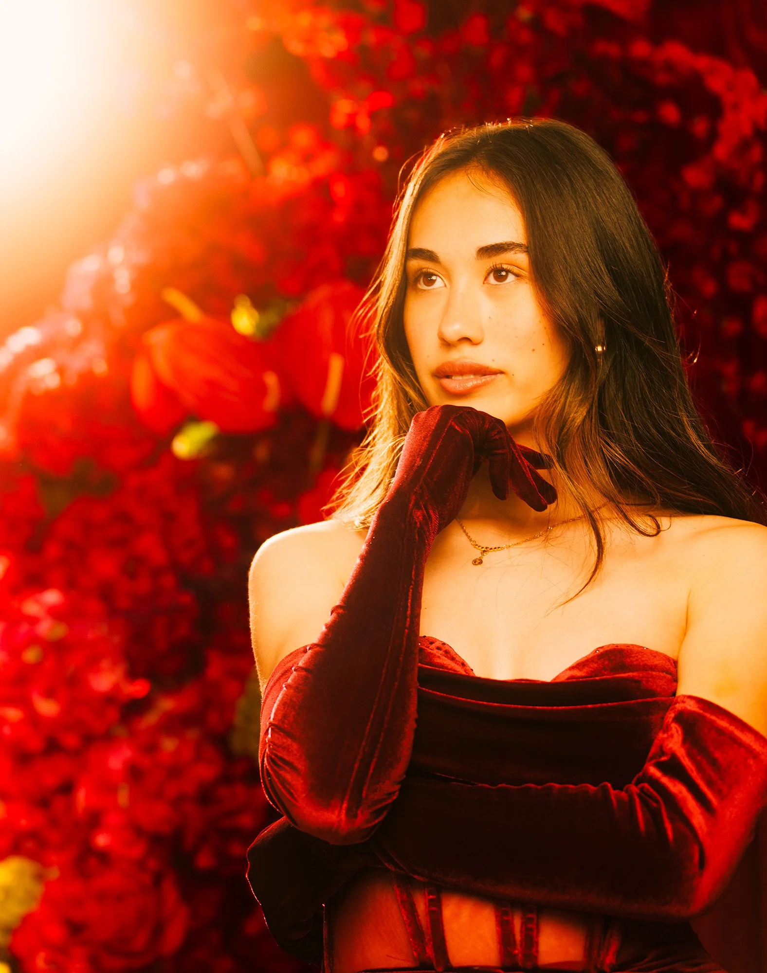 Editorial portrait of a woman in a red velvet dress and gloves, posed against a red floral backdrop with warm cinematic lighting and lens flare.