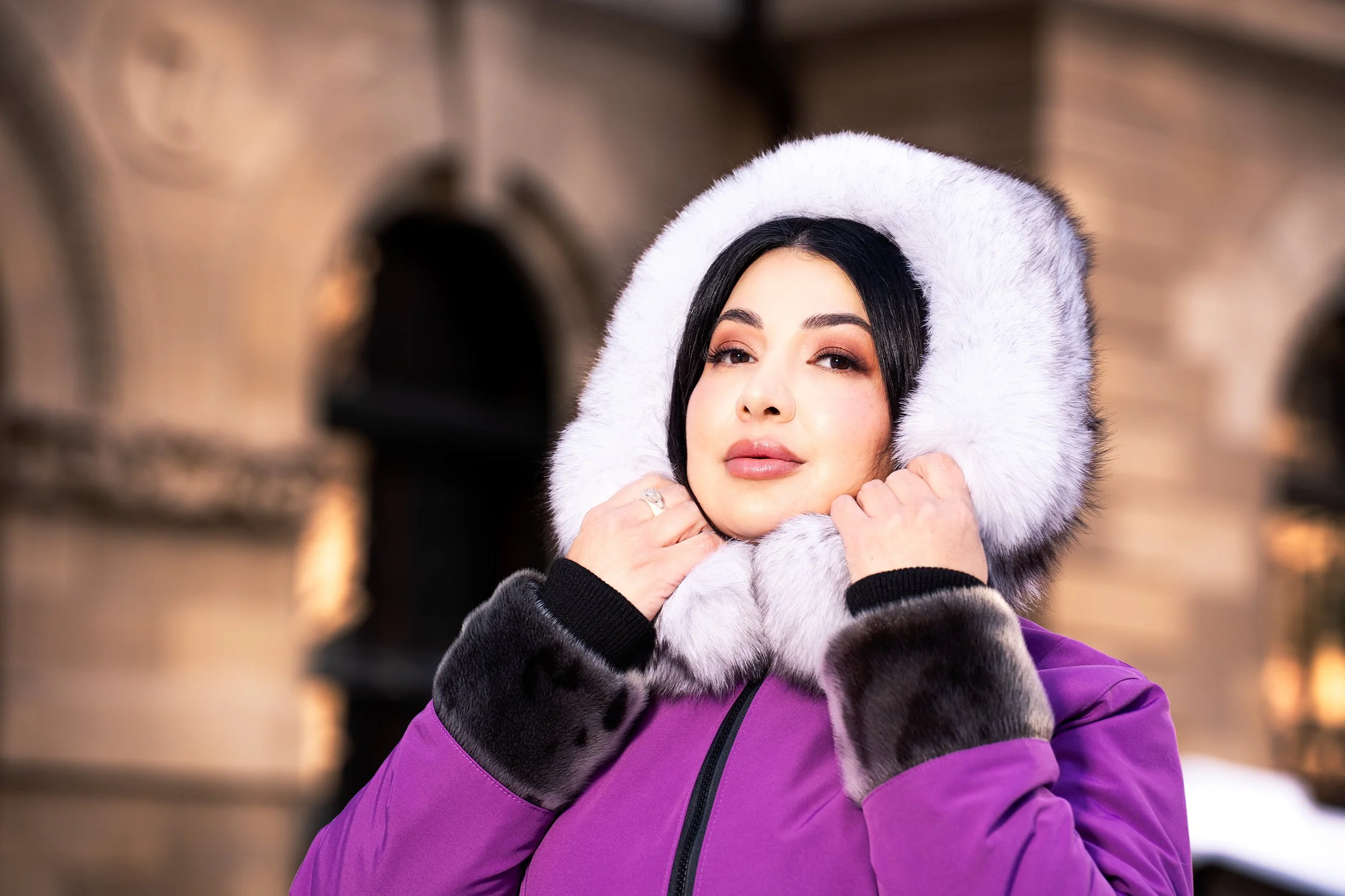 Elegant winter portrait of a woman in a purple coat with a fur-lined hood, softly lit against blurred stone architecture, highlighting warm skin tones and refined fashion styling.