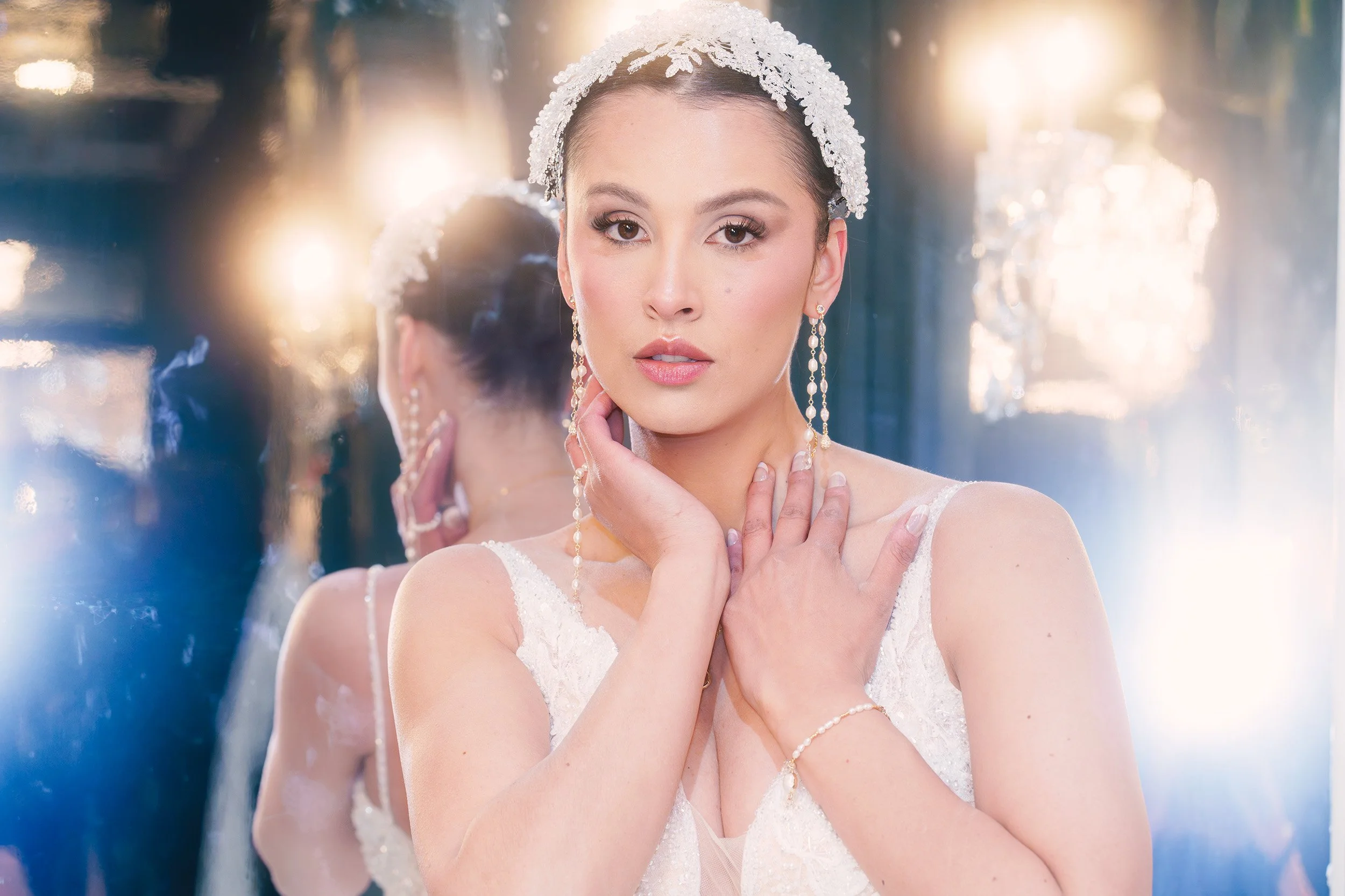 Cinematic bridal portrait of a woman in a white beaded gown and headpiece, posed in front of a mirror with glowing chandelier lights and blue-white lens flare.