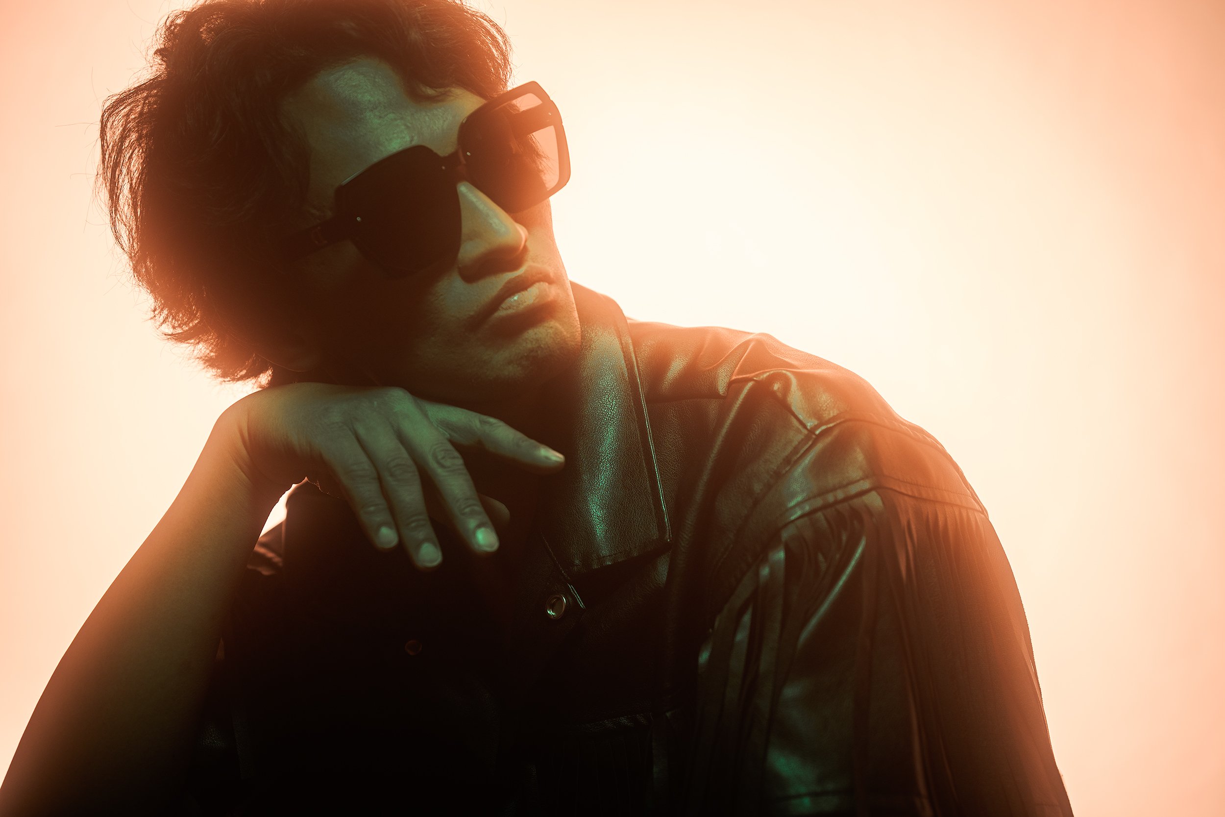Cinematic portrait of a man in a black leather jacket and oversized sunglasses, leaning with hand posed delicately, lit with dramatic teal and copper gradient lighting against a minimal studio background.