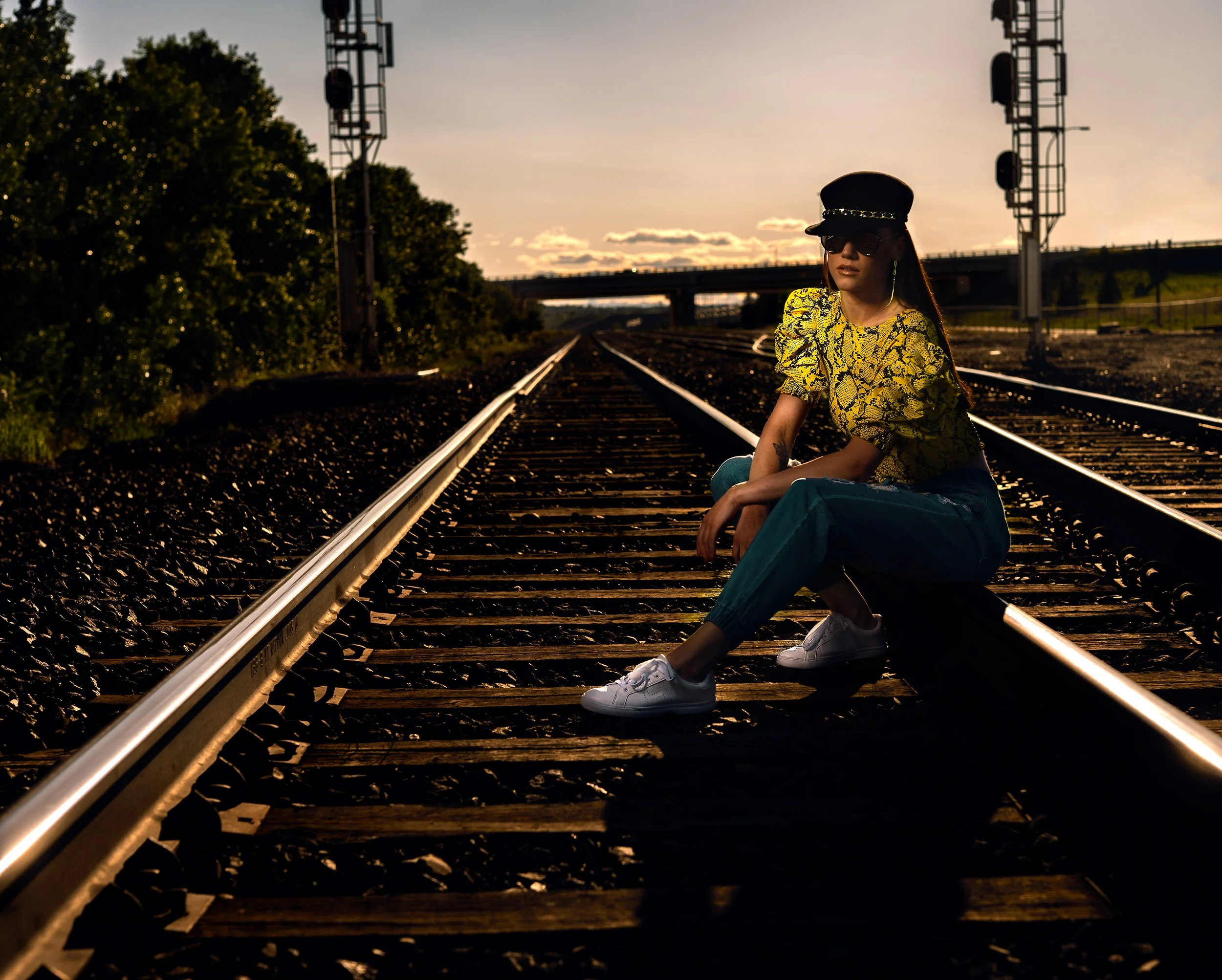 Fashion portrait of a woman sitting on railway tracks at sunset wearing a yellow patterned blouse and cap.