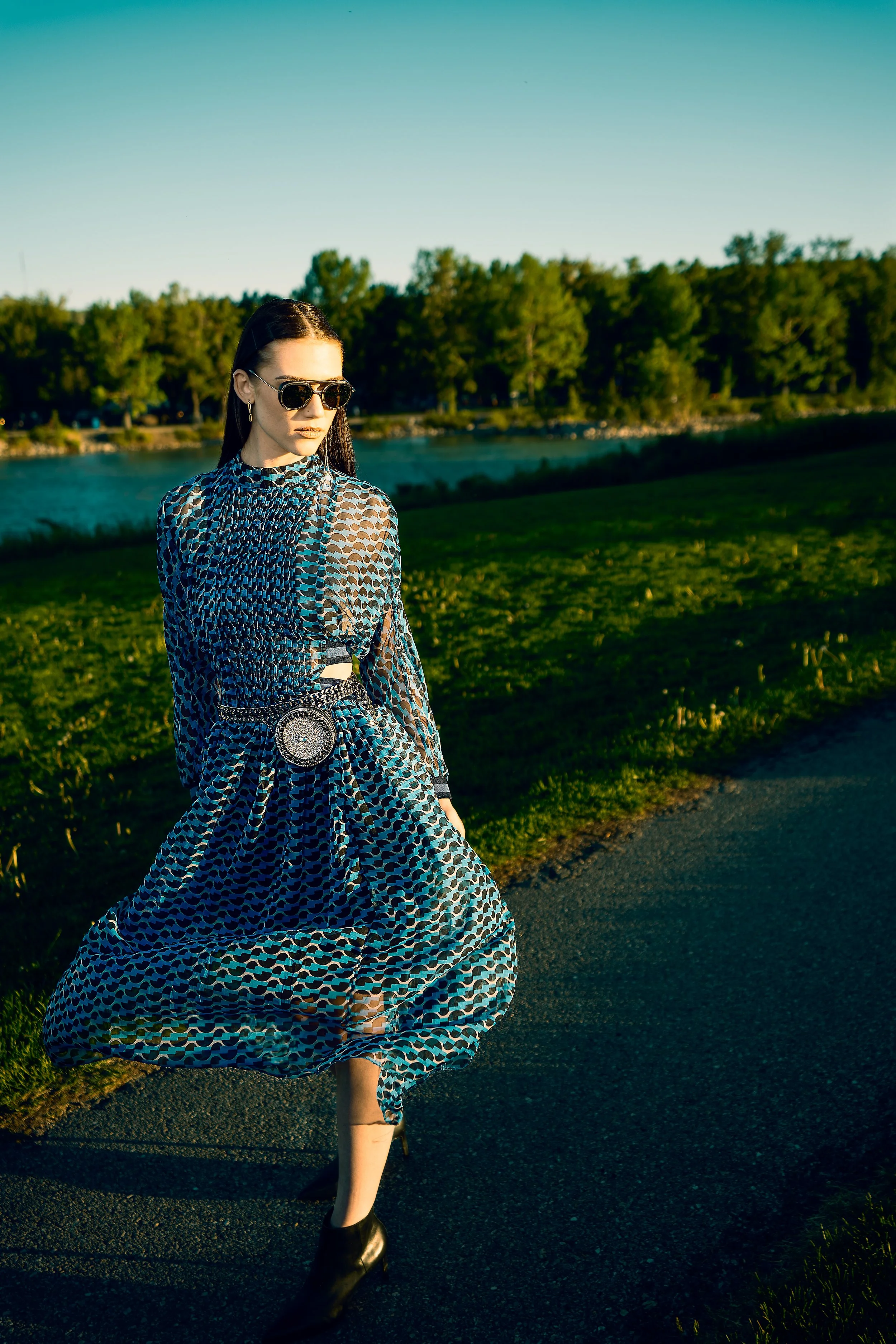 Editorial portrait photography of a woman in a patterned dress walking along a riverside path at golden hour, cinematic outdoor portrait session.
