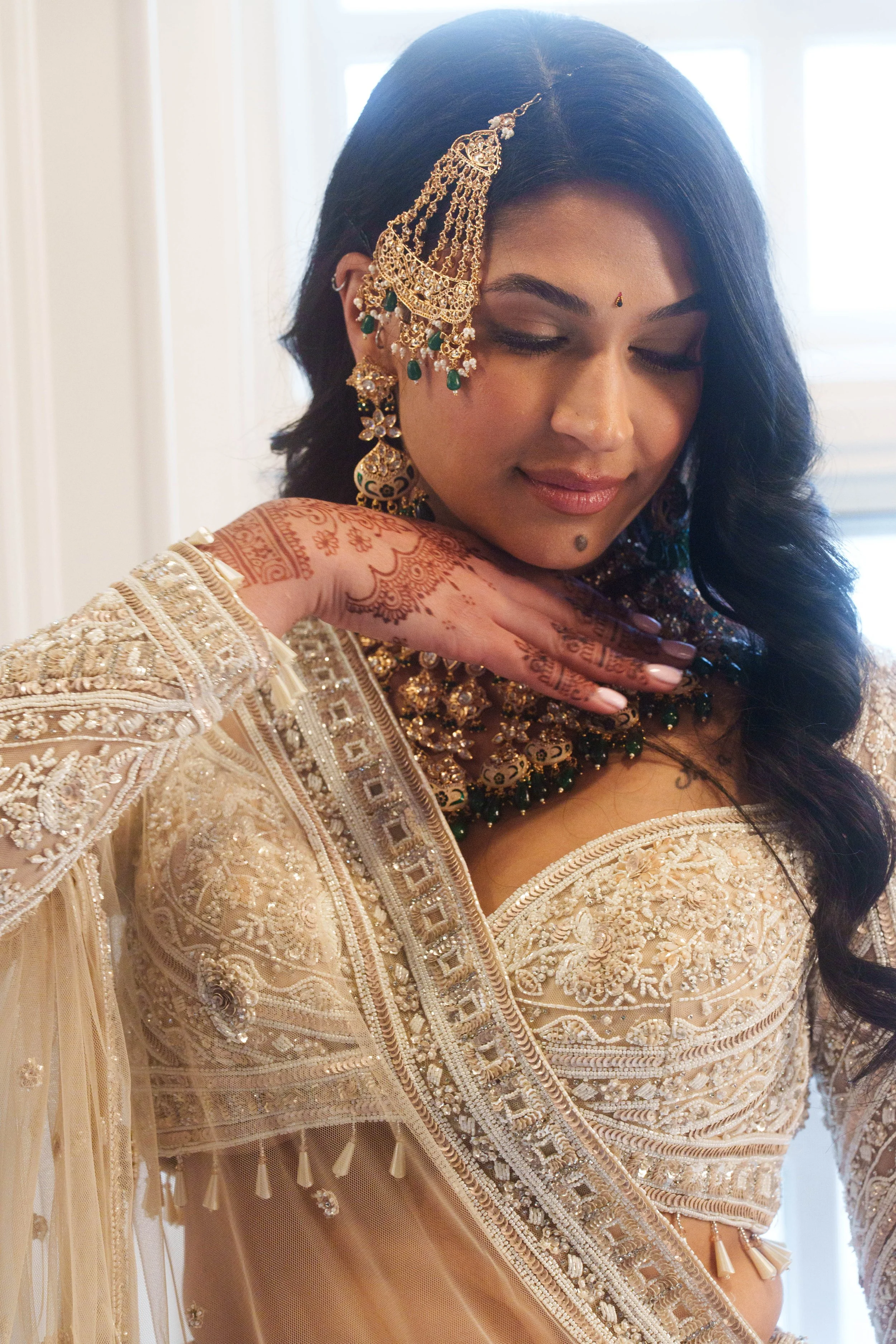 Close-up bridal portrait of a woman in ornate cream traditional attire, gold and emerald jewelry, and henna, posed with her hand beneath her chin in soft window light.