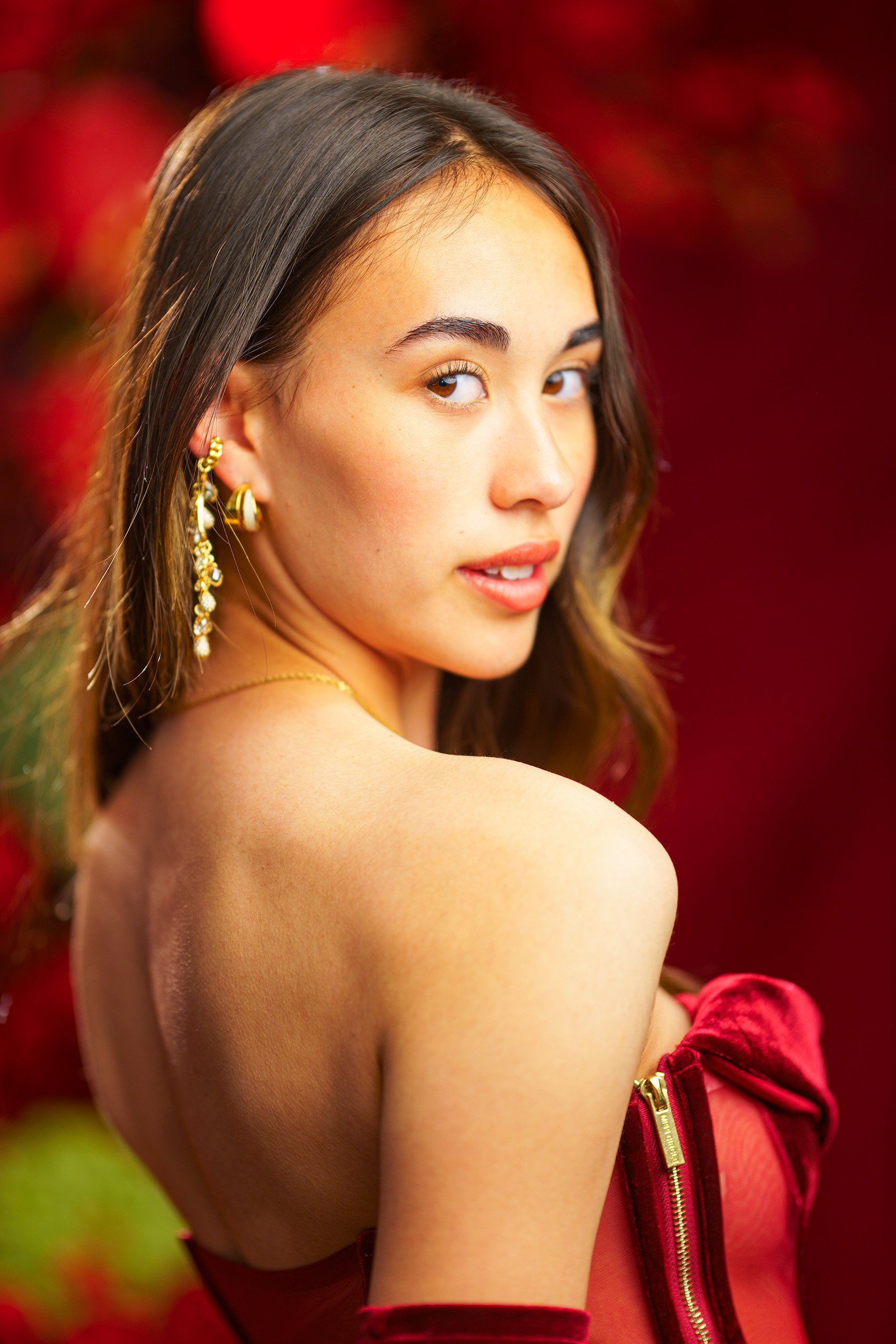 Close-up editorial portrait of a woman looking over her shoulder, wearing gold earrings and a red outfit against a soft red background.