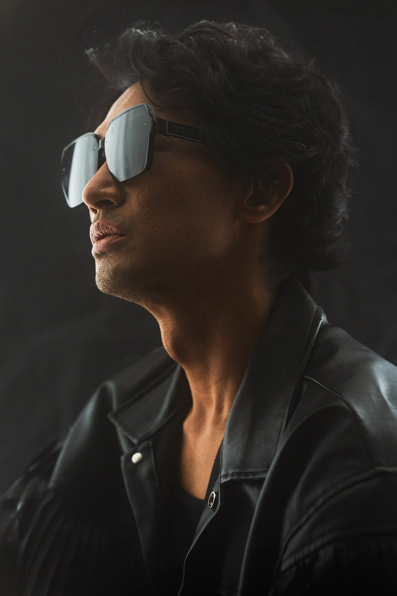 Professional fashion portrait of a man in black leather jacket and sunglasses, side profile with soft studio lighting and a dark blurred background.