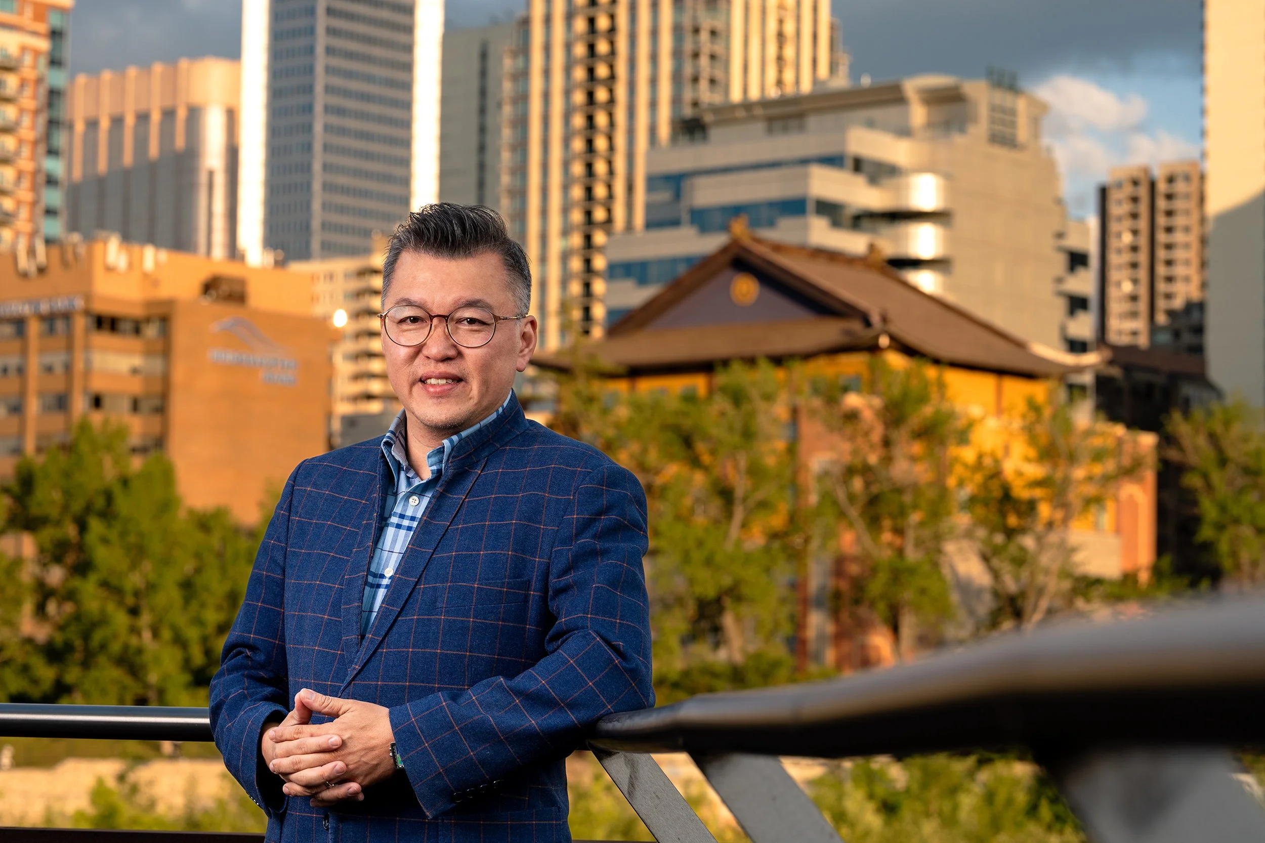 Golden hour portrait of a man in a blue blazer standing on a bridge, with leading lines from the railing guiding toward him against an urban skyline.