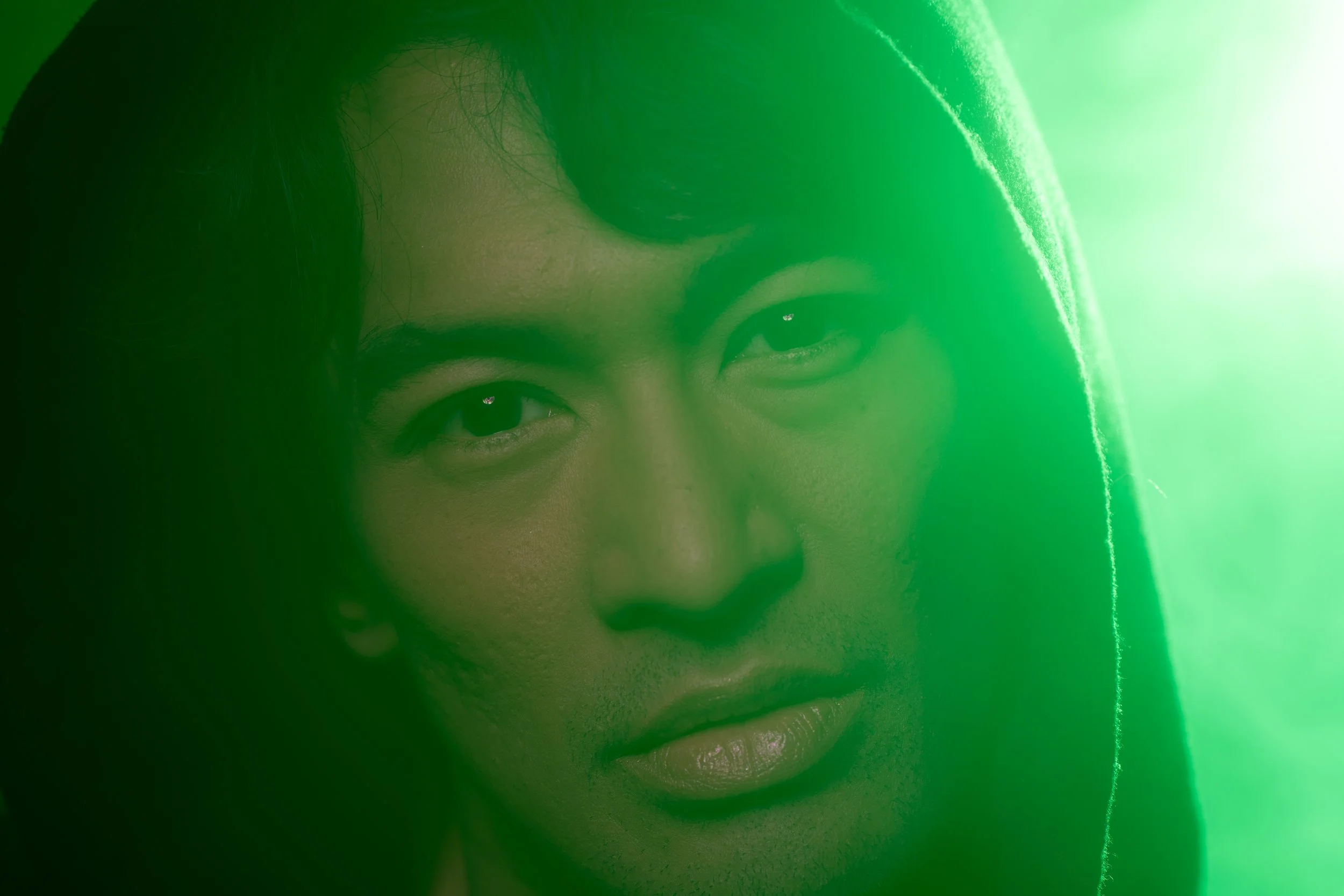 Close-up cinematic portrait of a man in a dark hood, partially obscured by green light flare, looking directly at the camera with an intense expression.