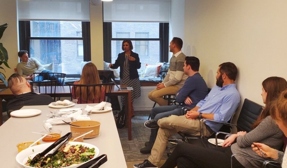 A woman standing and speaking to a group of seven people in a room with large windows, some seated in chairs and one lounging on a window seat.