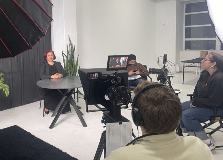 A woman sitting at a round table, being filmed in a studio with a camera, crew members, and lighting equipment, with a large plant and white walls in the background.