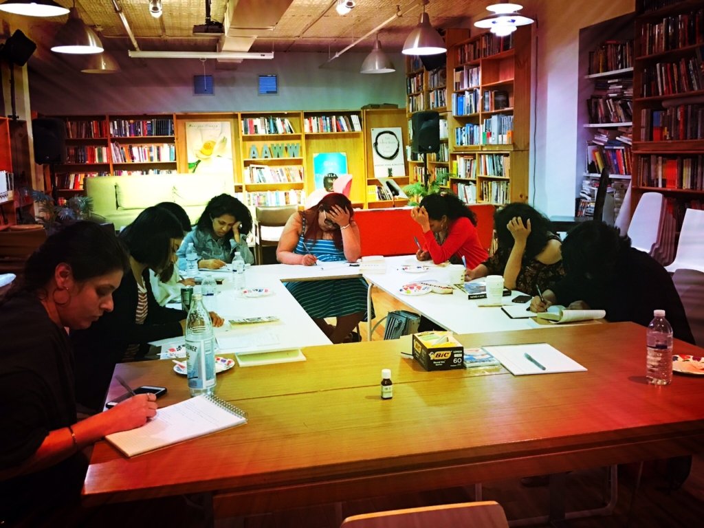 Six women seated around a large rectangular table in a cozy library or bookstore, engaged in writing or reading. The room has wooden bookshelves filled with books, a green sofa, colorful artwork, and soft overhead lighting.