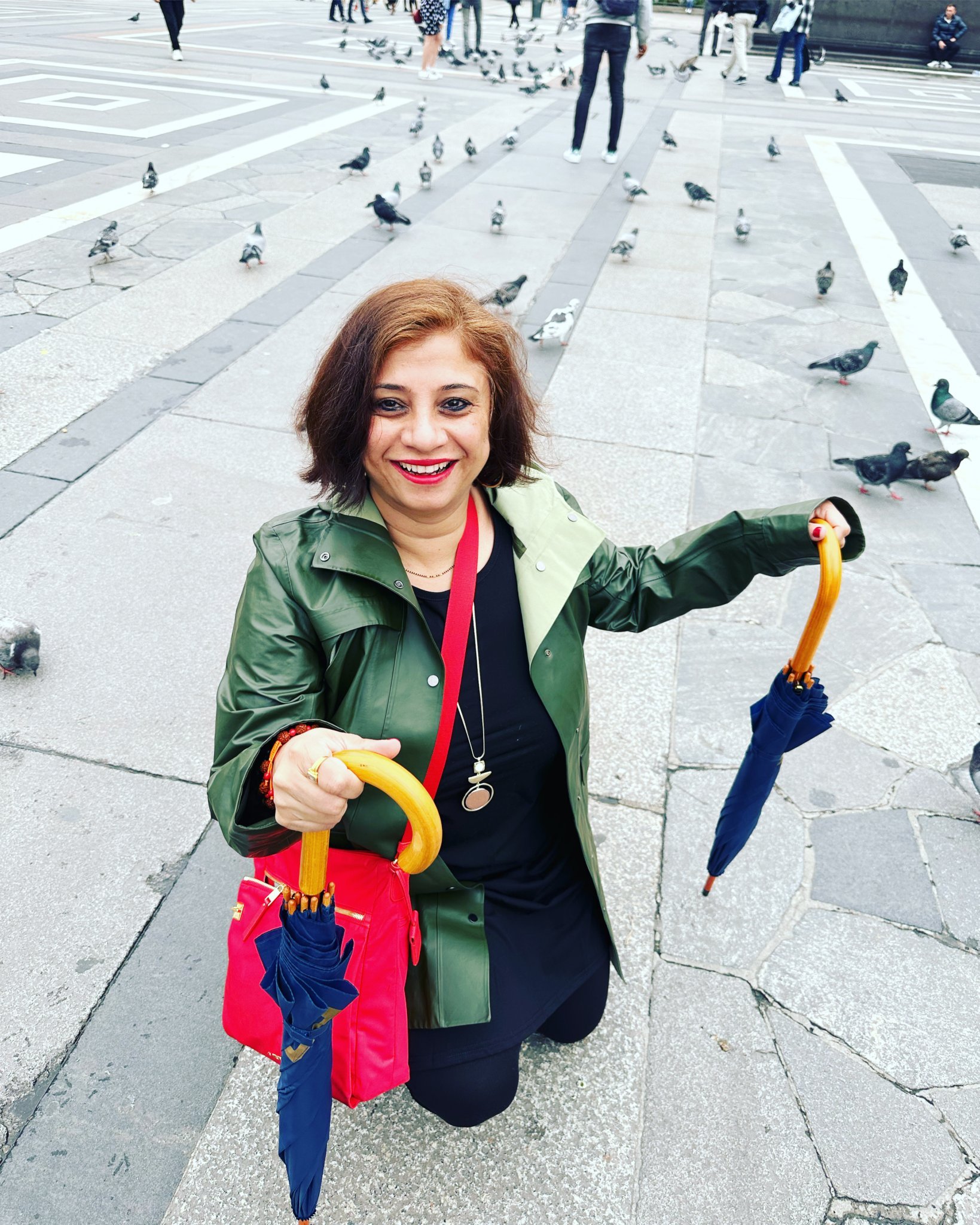 A woman with shoulder-length brown hair, red lipstick, and a green jacket crouching on a city sidewalk, smiling at the camera, holding two closed umbrellas with yellow handles.