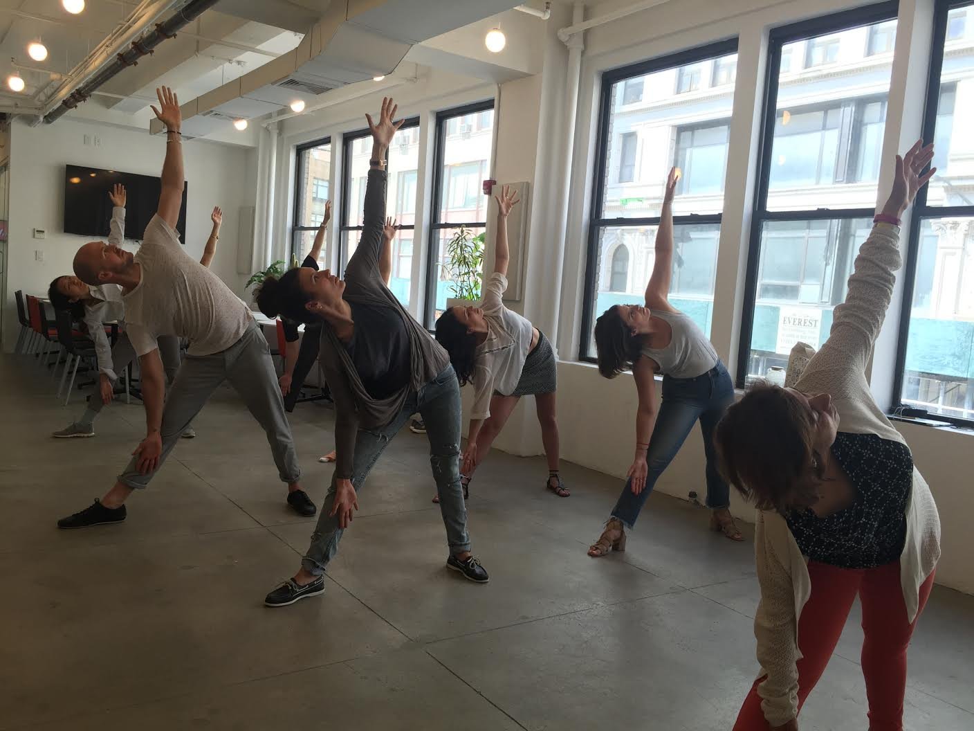 Group of people in a yoga class, performing a side stretch pose with one arm raised overhead, in a bright room with large windows.