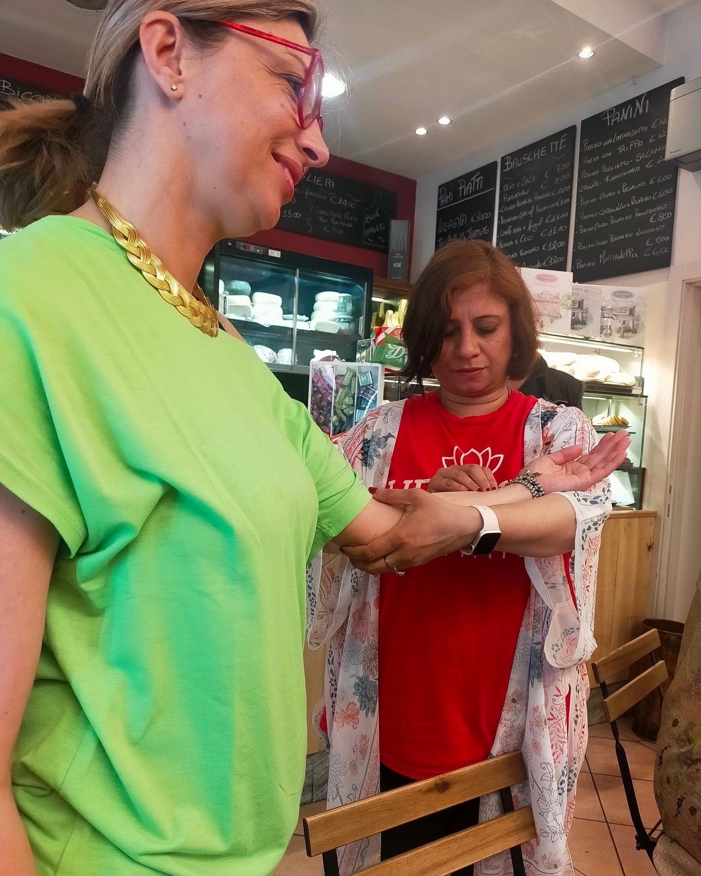 Two women in a cafe, one wearing a bright green shirt and glasses, holding out her arm, while the other woman in a red shirt and floral cover-up examines her wrist wearing a smartwatch, with menu boards in the background.