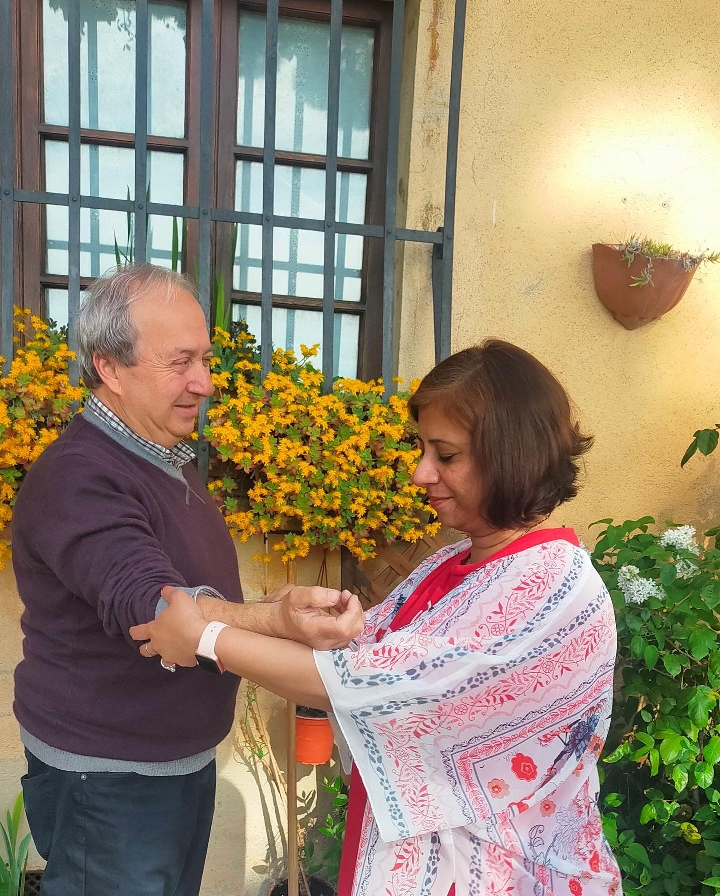 A man and woman standing outdoors in front of a yellow wall and greenery, holding hands and looking at each other, appearing to share a tender moment.
