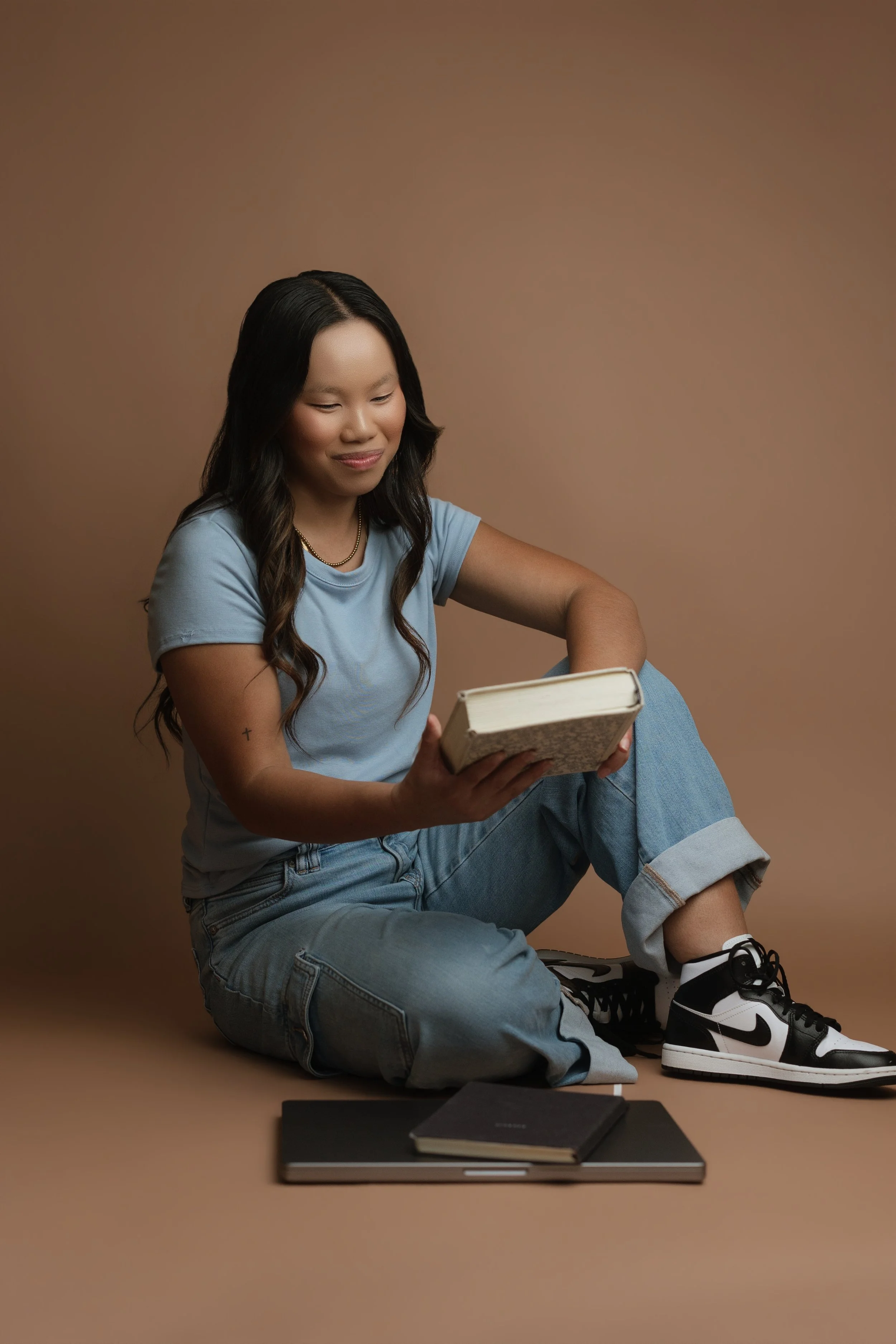 A young woman with long dark hair, wearing a light blue T-shirt, ripped jeans, and black and white sneakers, is sitting on the floor with a beige background, reading a book.
