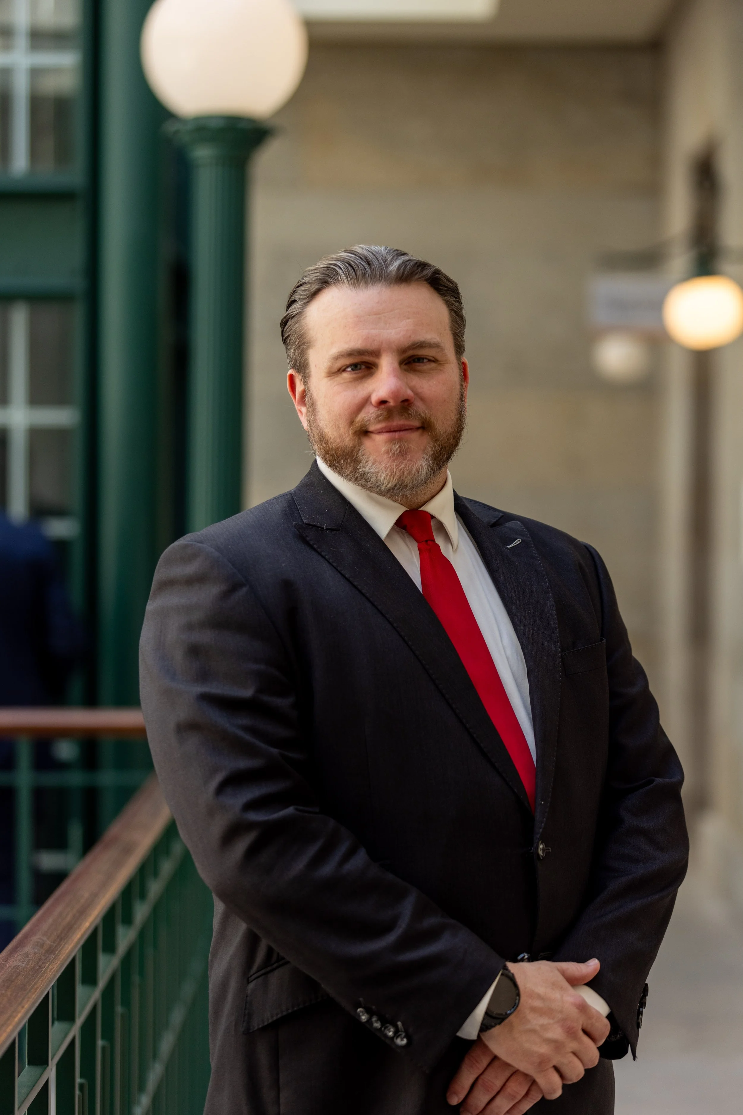 A man in a dark suit and red tie standing outside near a green railing and white lamp post.