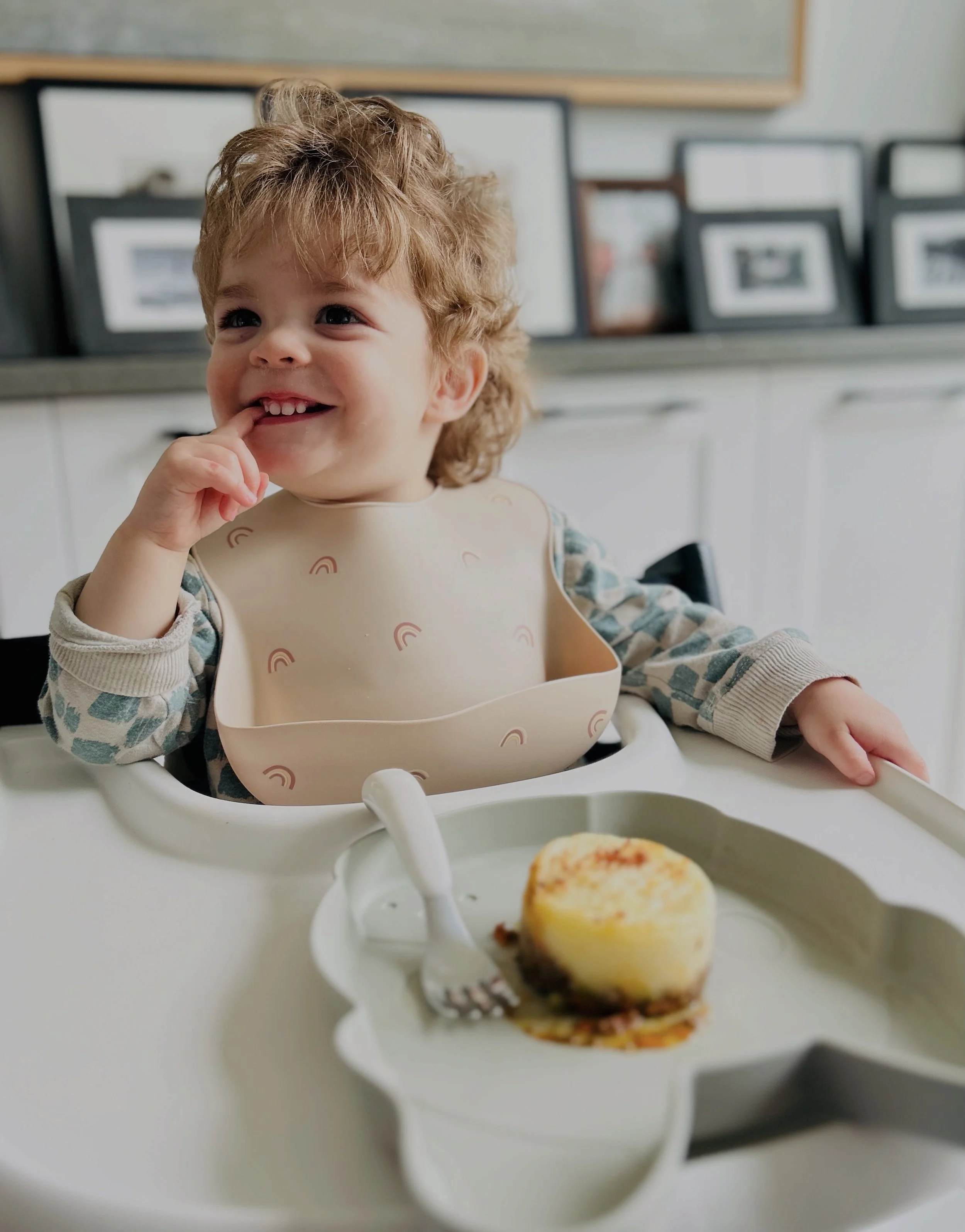 A happy young child with curly hair, sitting in a high chair and smiling, wearing a bib and long-sleeved shirt, with a plate of mini cottage pie in front of them on a tray, in a dining room with framed photographs in the background.