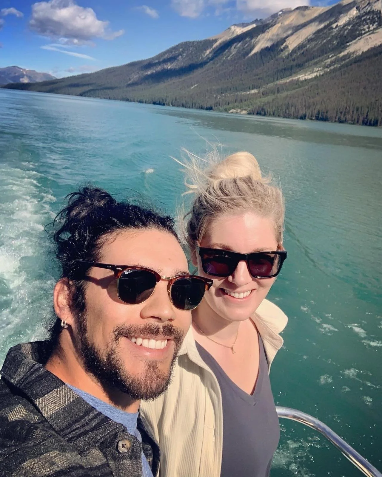 A smiling man and woman wearing sunglasses taking a selfie on a boat with a scenic lake and mountains in the background.