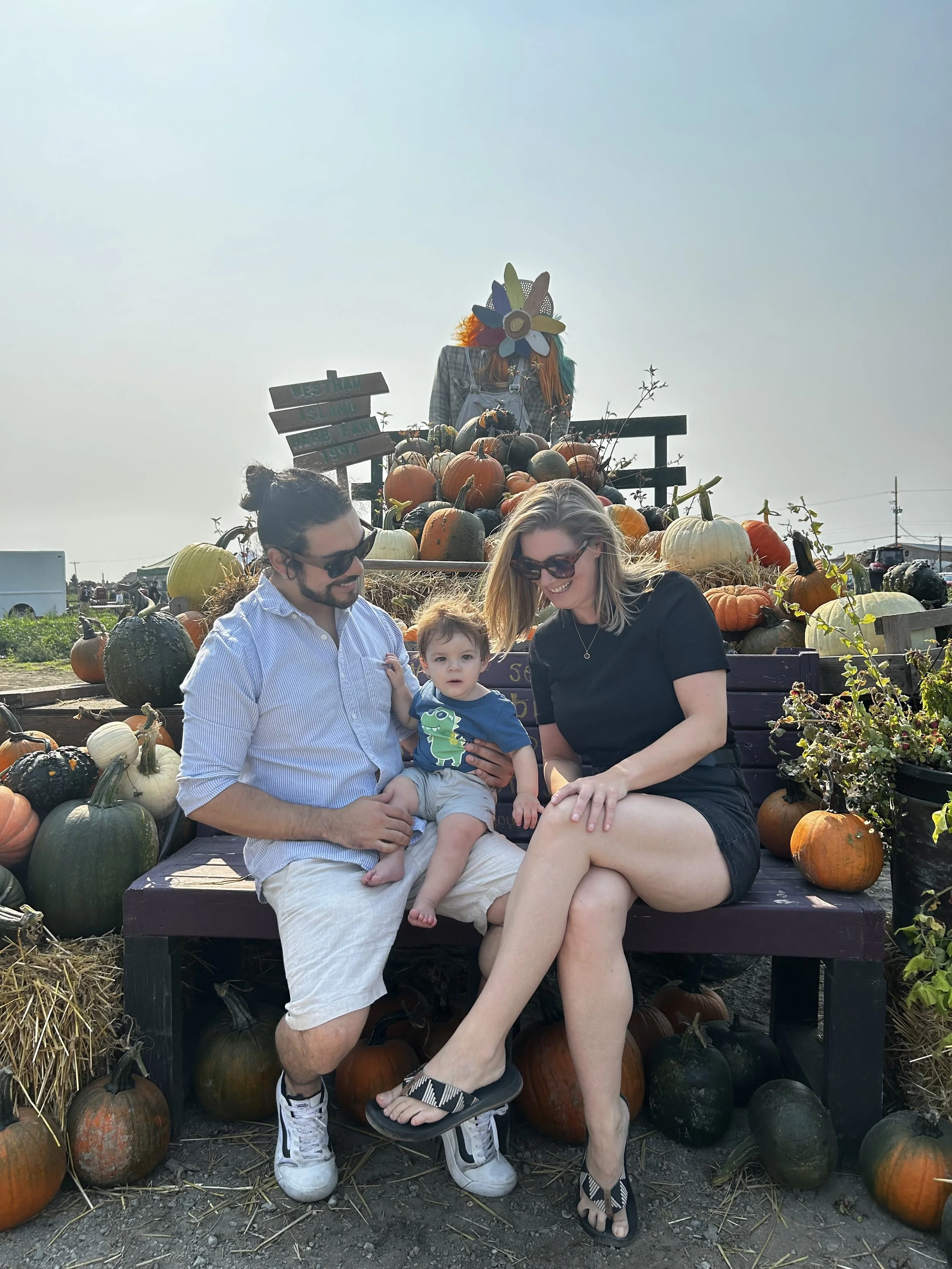 A family of three sitting on a bench at a pumpkin patch, with a variety of pumpkins around them, and a scarecrow with a colorful mask in the background.