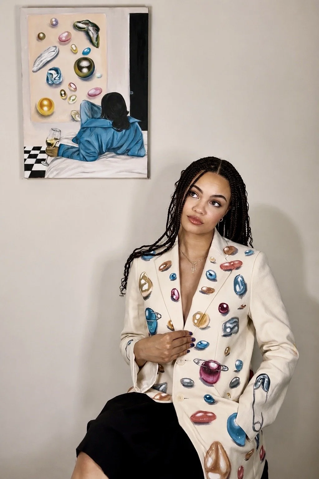 A woman with braided hair wearing a cream-colored blazer decorated with colorful 3D gemstones, sitting with one leg crossed and her right hand on her chest, posing in front of a plain wall and a contemporary painting of a person with long dark hair wearing a blue shirt, lying in bed and looking at an abstract art piece.