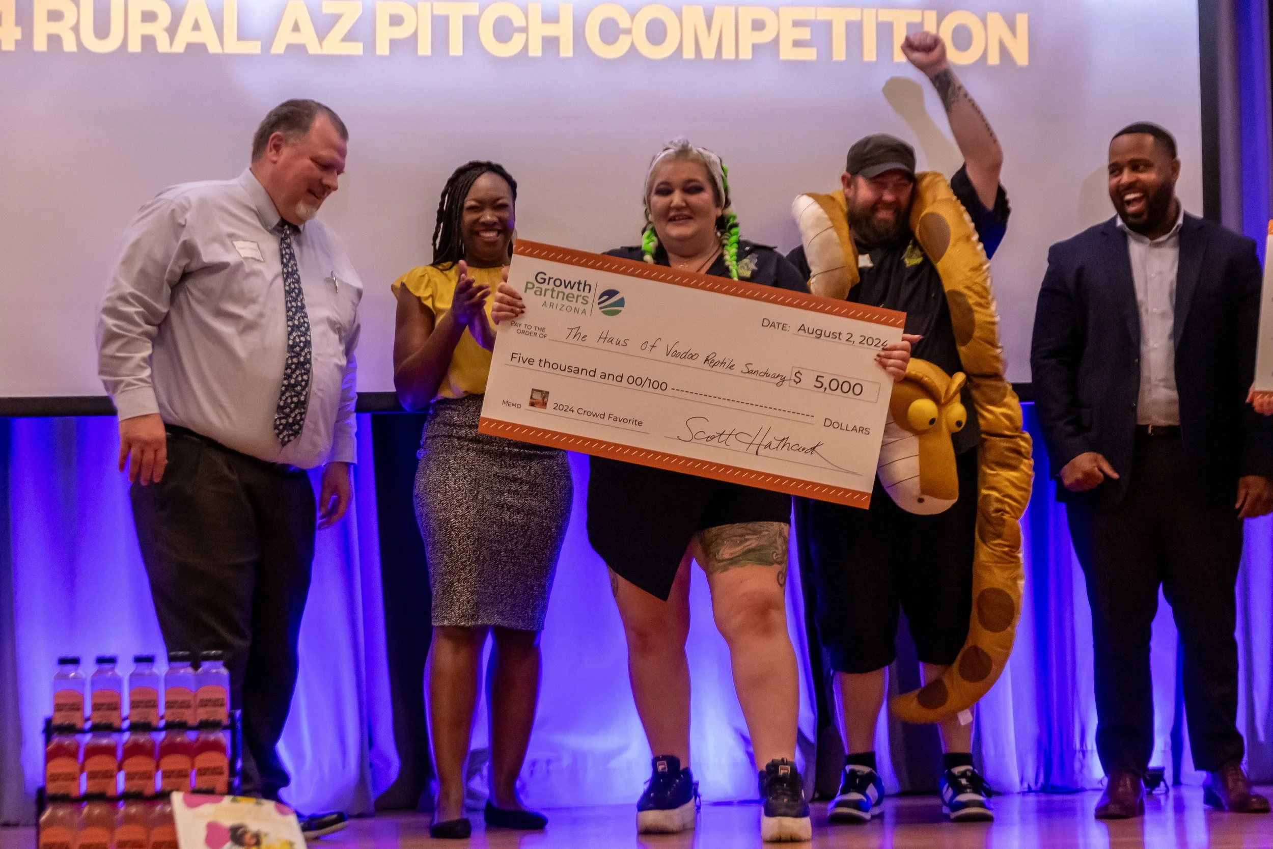 Group of people on stage holding a large ceremonial check for $5,000 made out to the Haus of Voodoo Reptile Sanctuary, with a backdrop reading 'RURAL AZ PITCH COMPETITION'. The group includes a woman with braids holding the check, a man in a yellow snake costume, and others dressed in business casual or colorful attire, celebrating their win.