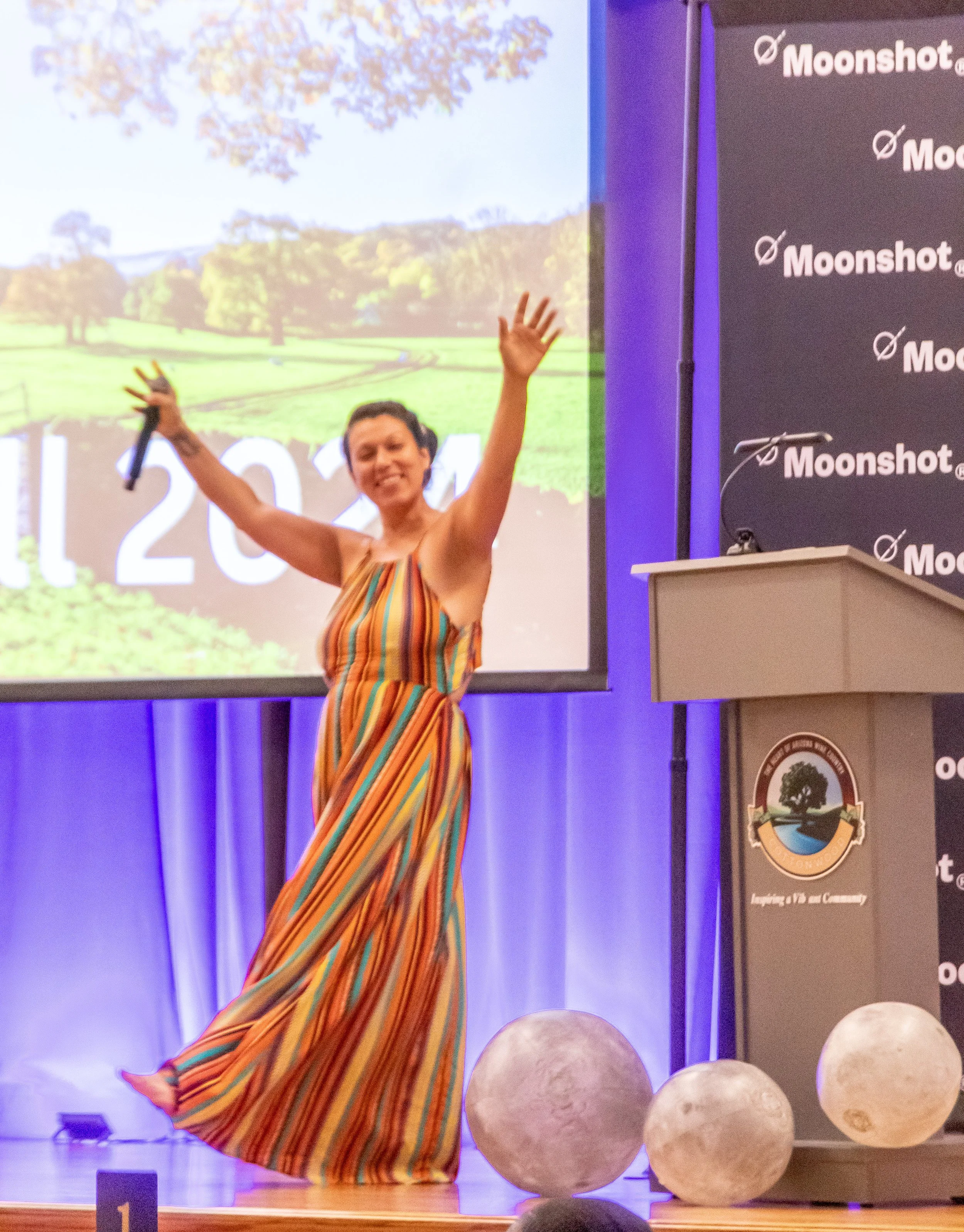 A woman in a colorful striped dress with arms raised, smiling, standing on a stage in front of a large screen and panel with Moonshot branding, with moon models on the floor in front.