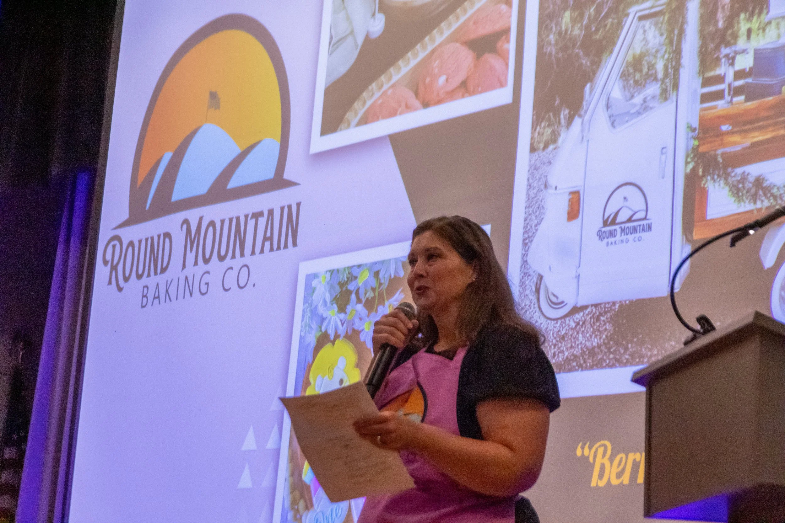 A woman giving a presentation on stage with a large screen behind her displaying the logo of Round Mountain Baking Co., which features a mountain and sunrise, along with images of baked goods and a delivery truck with the same logo.