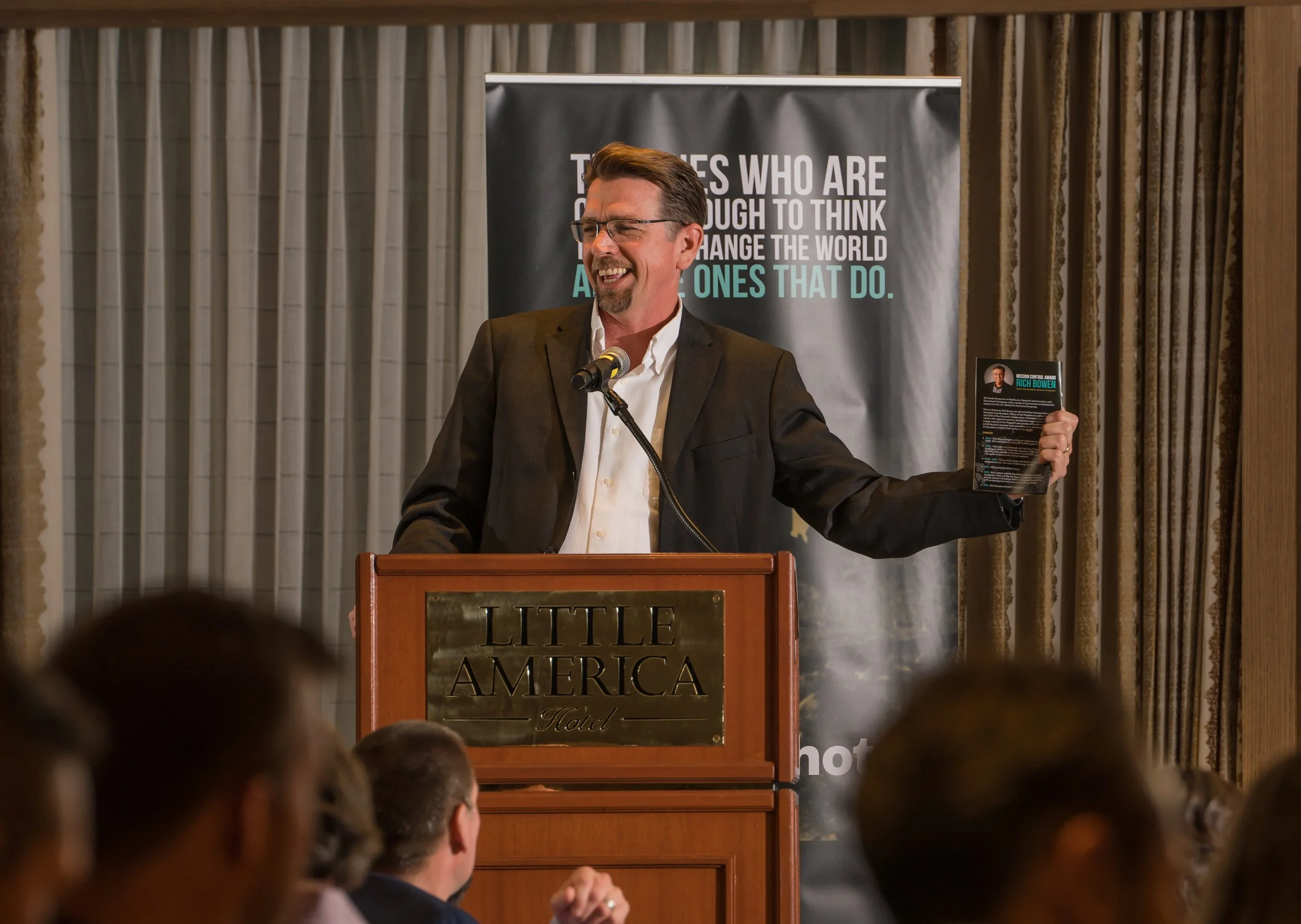 A man in glasses and a dark suit speaking at a podium with a sign that reads "LITTLE AMERICA Hotel". He is holding a pamphlet and smiling, with an audience in front of him. Behind him is a black banner with inspirational text.