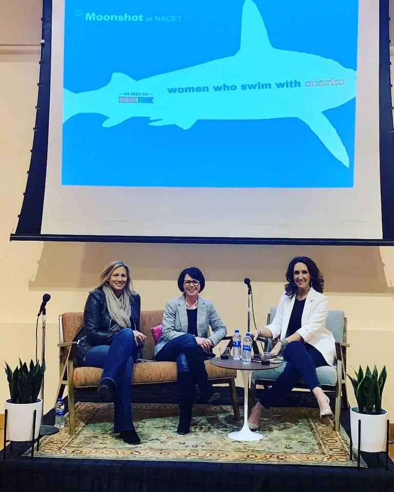 Three women sitting on a sofa in front of a presentation screen, with a title about women who swim with sharks.